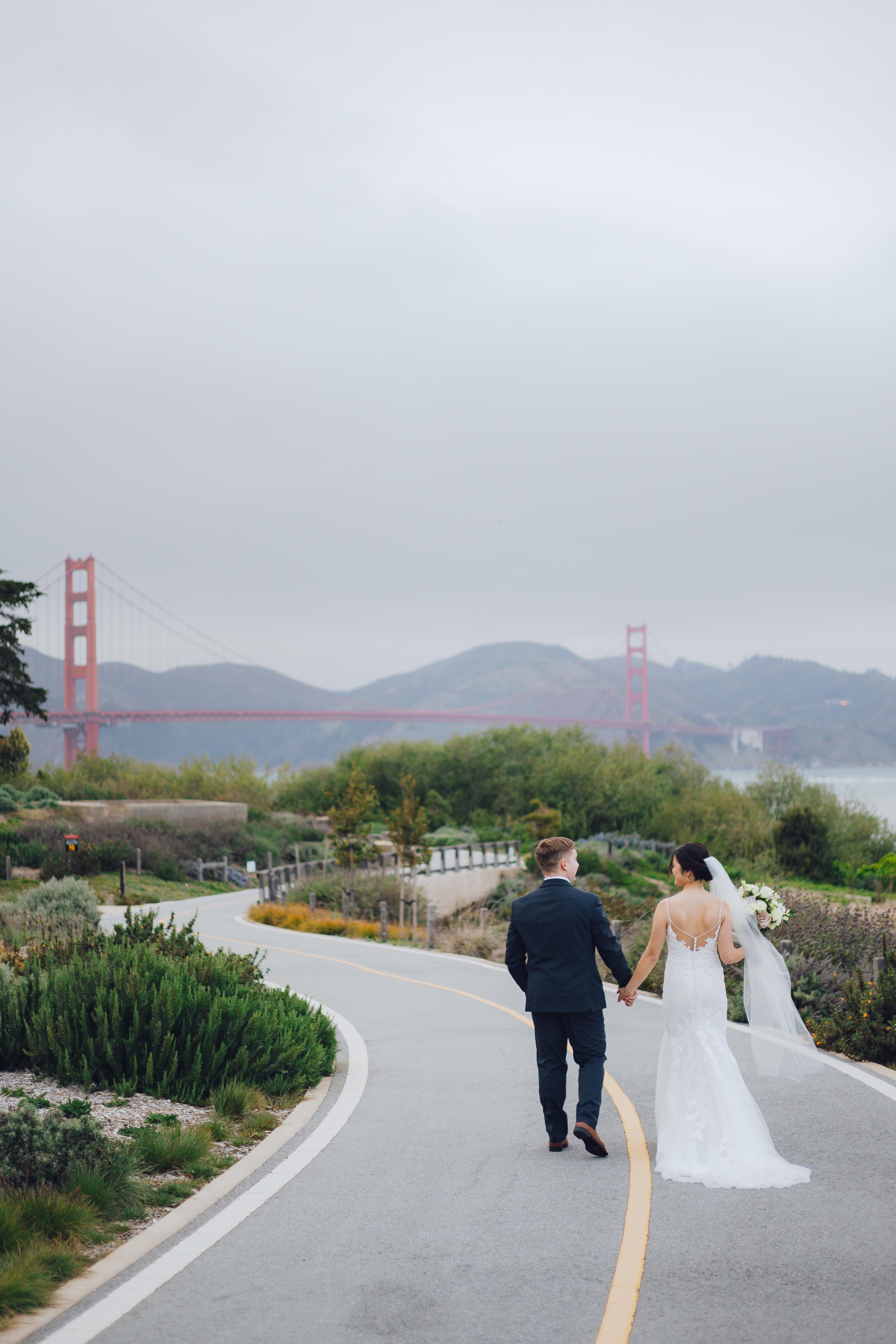 Bride in white dress holding bouquet and groom in suit holding hands walking on a curved path with the Golden Gate Bridge in the background.