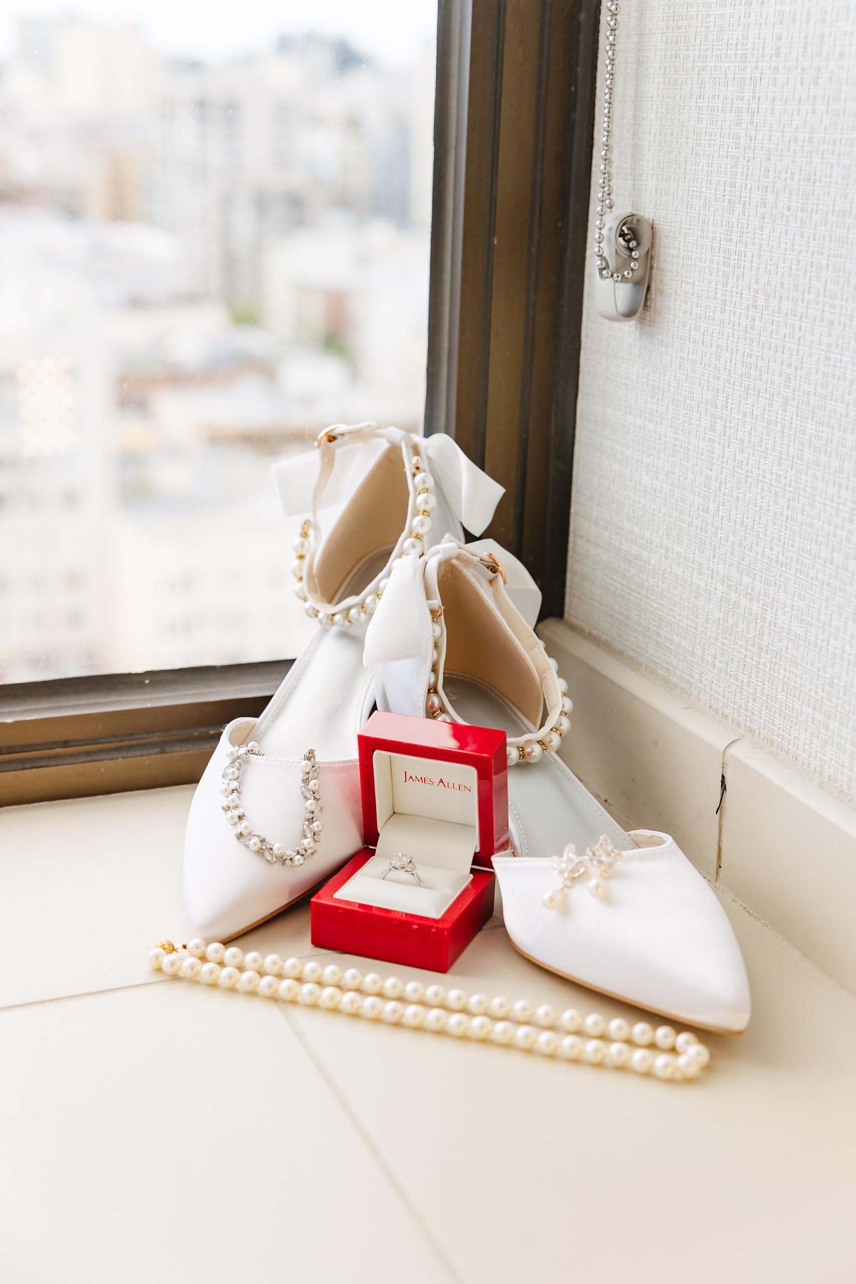 White bridal shoes decorated with pearls, a pearl necklace, earrings, and an engagement ring in a red James Allen box near a window.