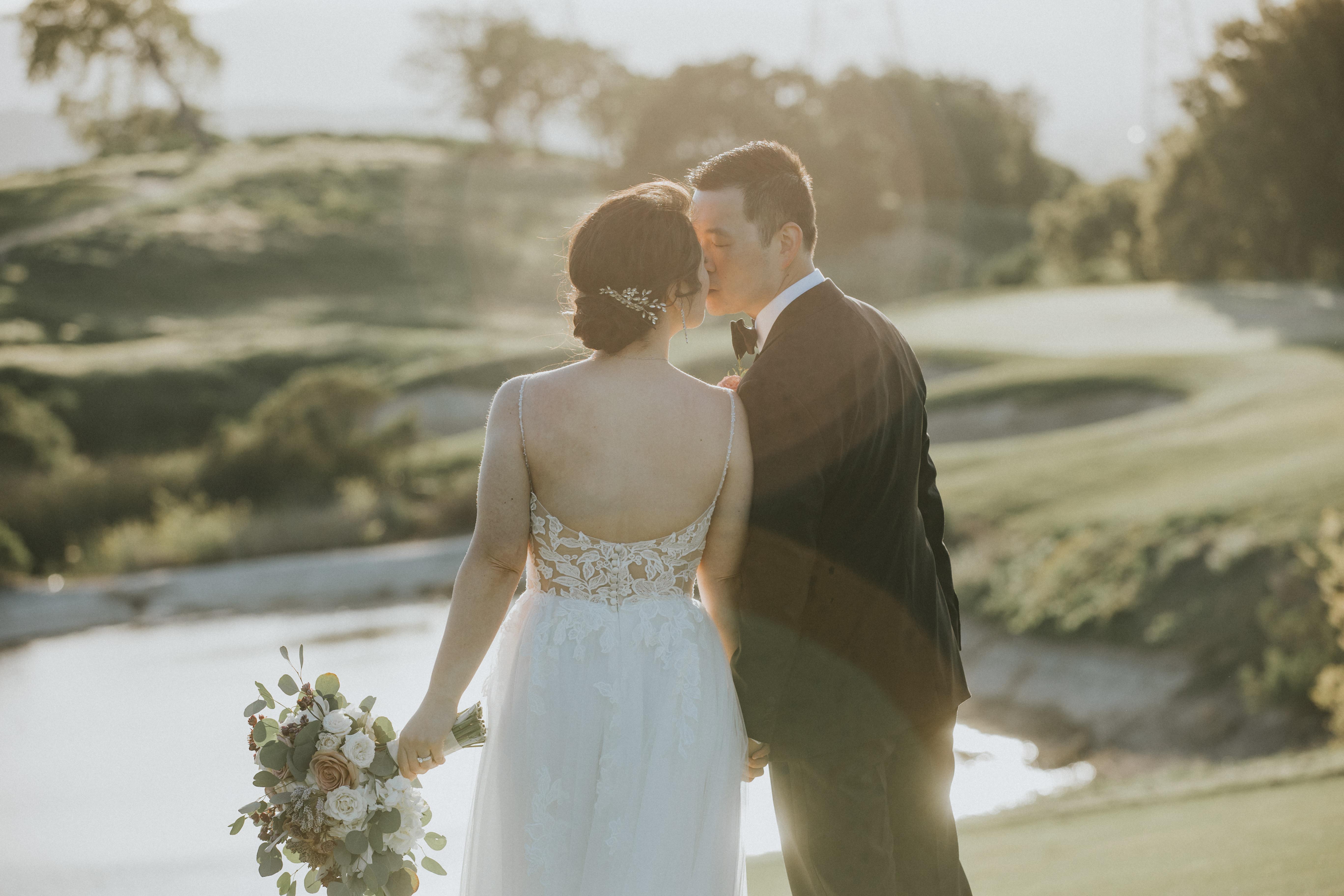 Bride and groom holding hands and kissing outdoors near a body of water during sunset.