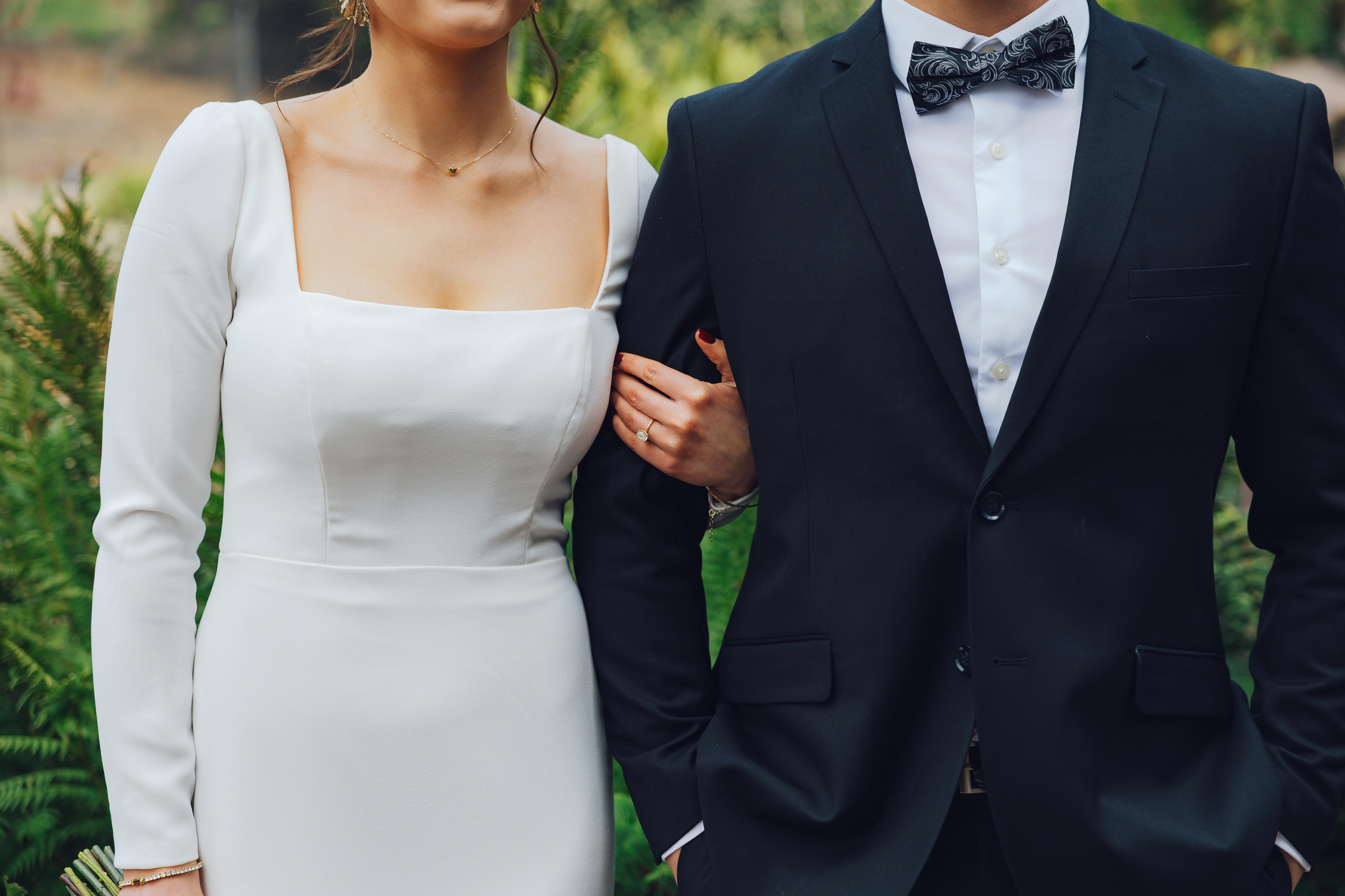 Woman in a white long-sleeve dress holding arm of a man wearing a black suit and patterned bow tie.
