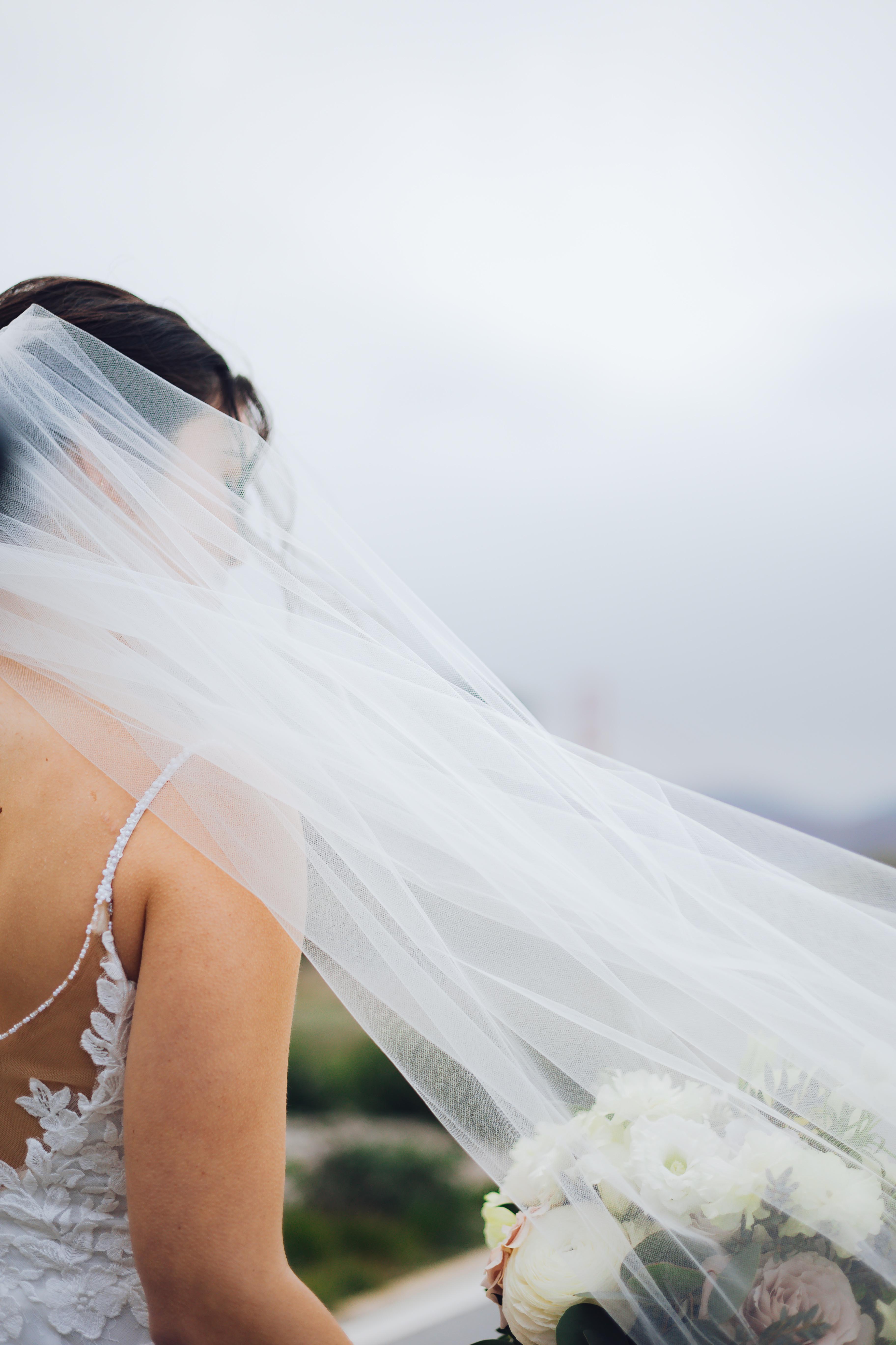 Bride in a white lace dress with a long veil blowing in the wind, holding a bouquet of white and pale pink flowers.
