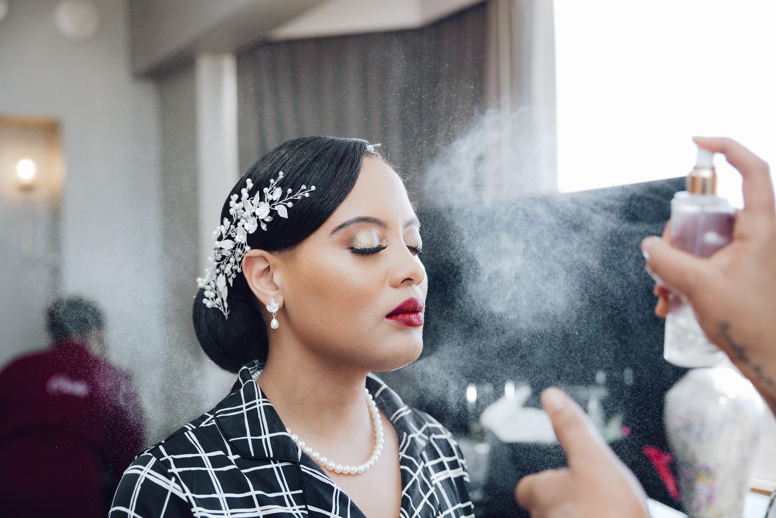 Woman with closed eyes wearing pearl necklace and earrings, having makeup setting spray misted on her face.