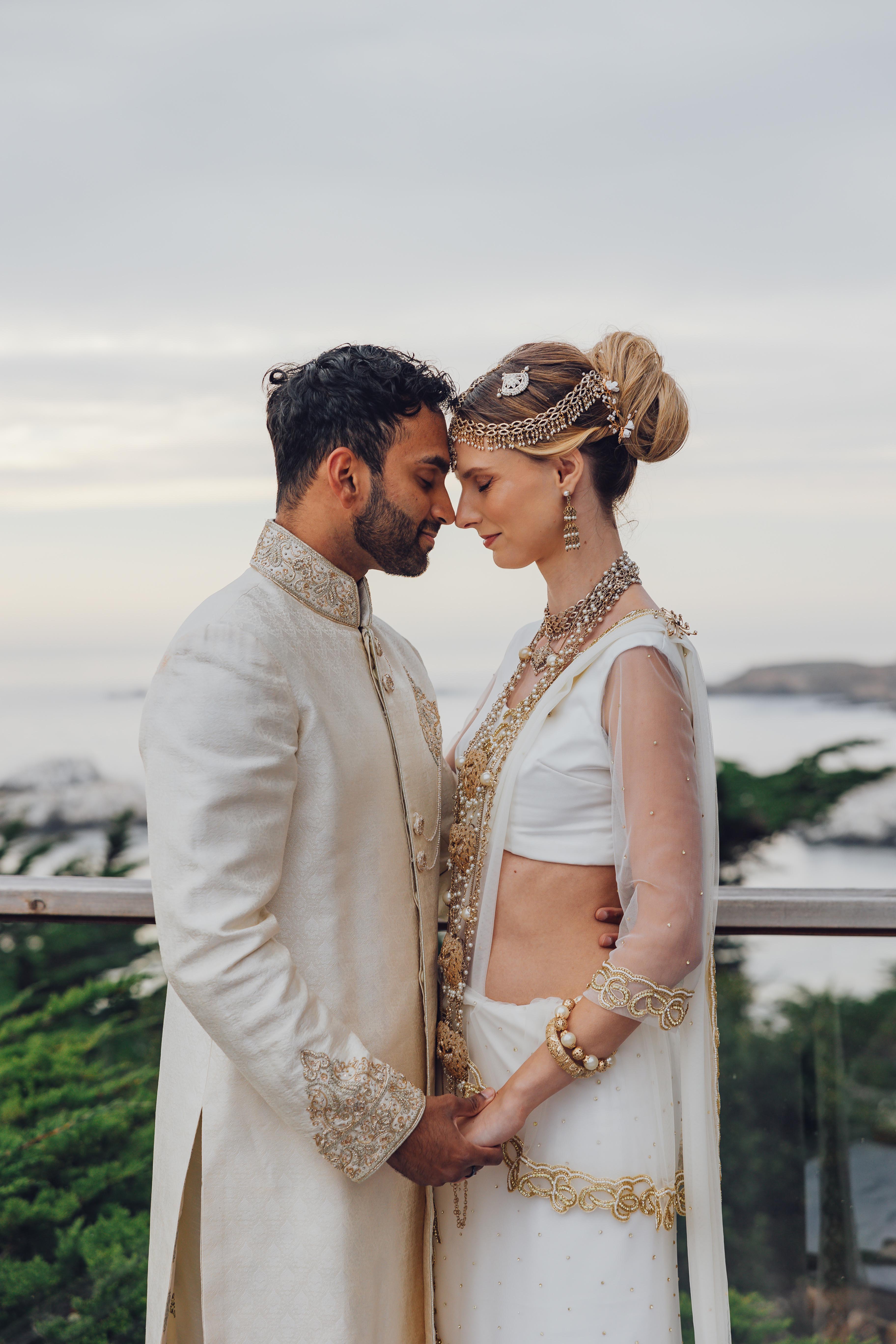 Bride and groom in traditional South Asian attire holding hands and touching foreheads outdoors near greenery and water.