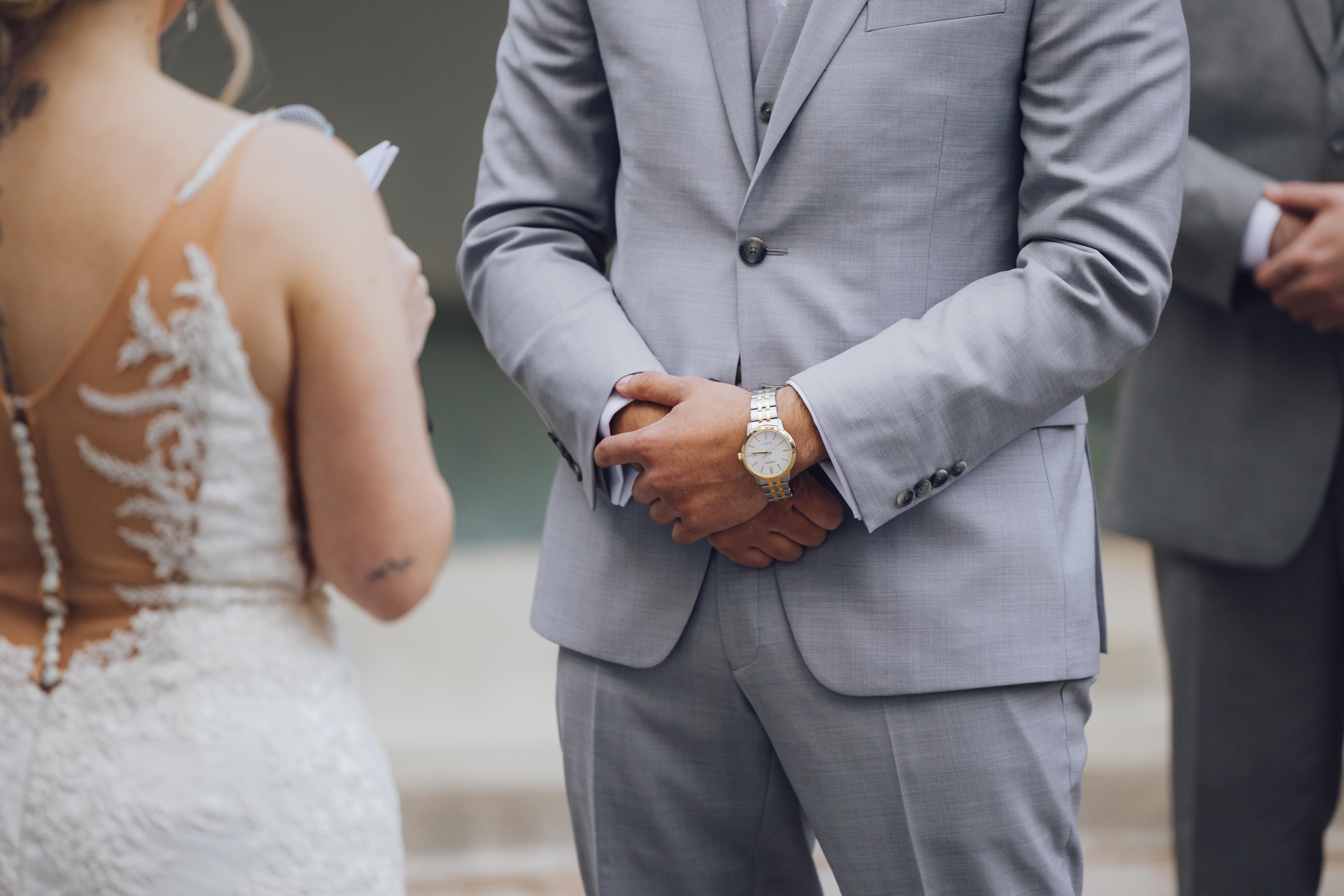Man in a light gray suit with his hands clasped in front wearing a wristwatch during a wedding ceremony, facing a woman in a white lace wedding dress.