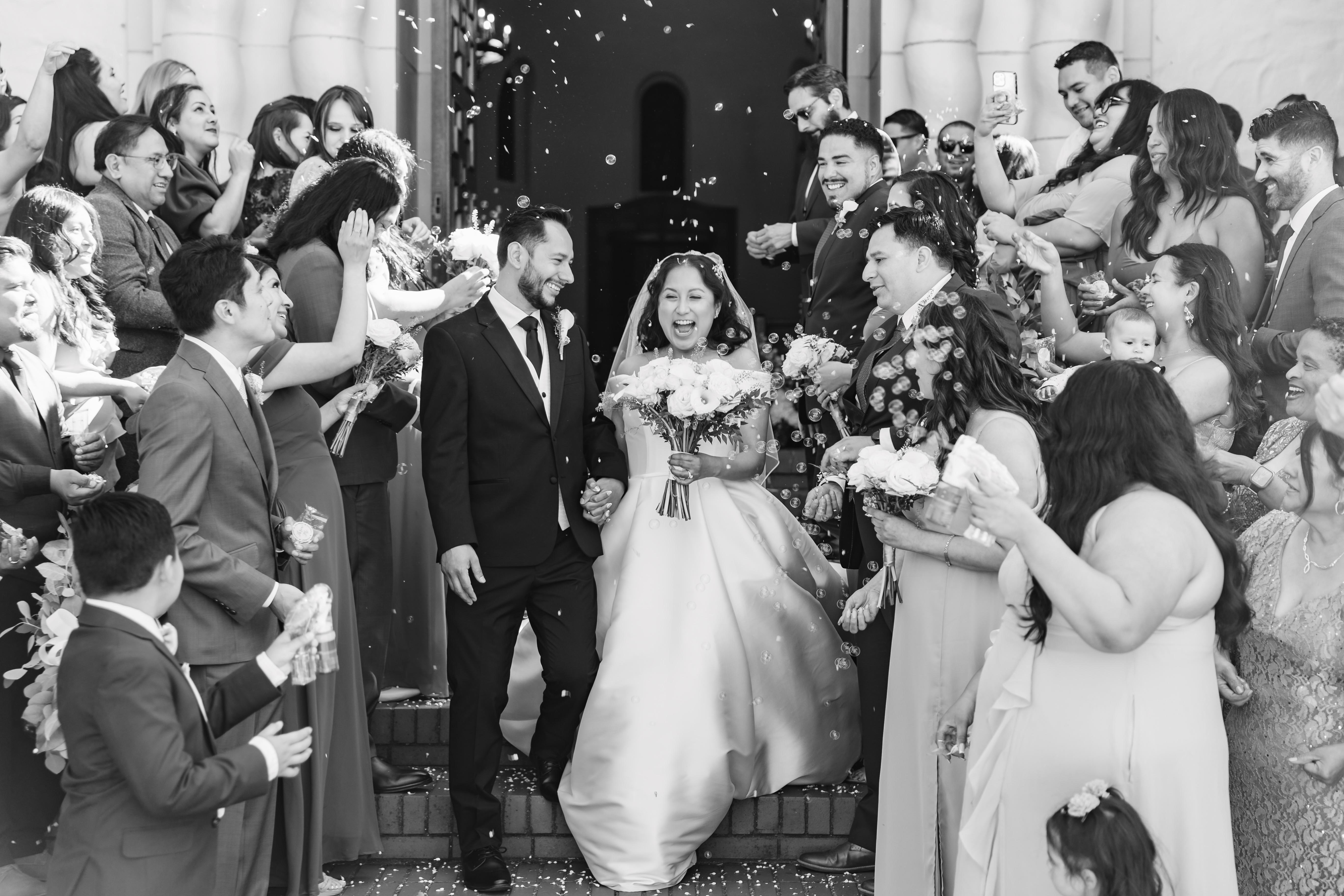 Bride and groom smiling and holding hands while guests surround them blowing bubbles and celebrating on stairs.