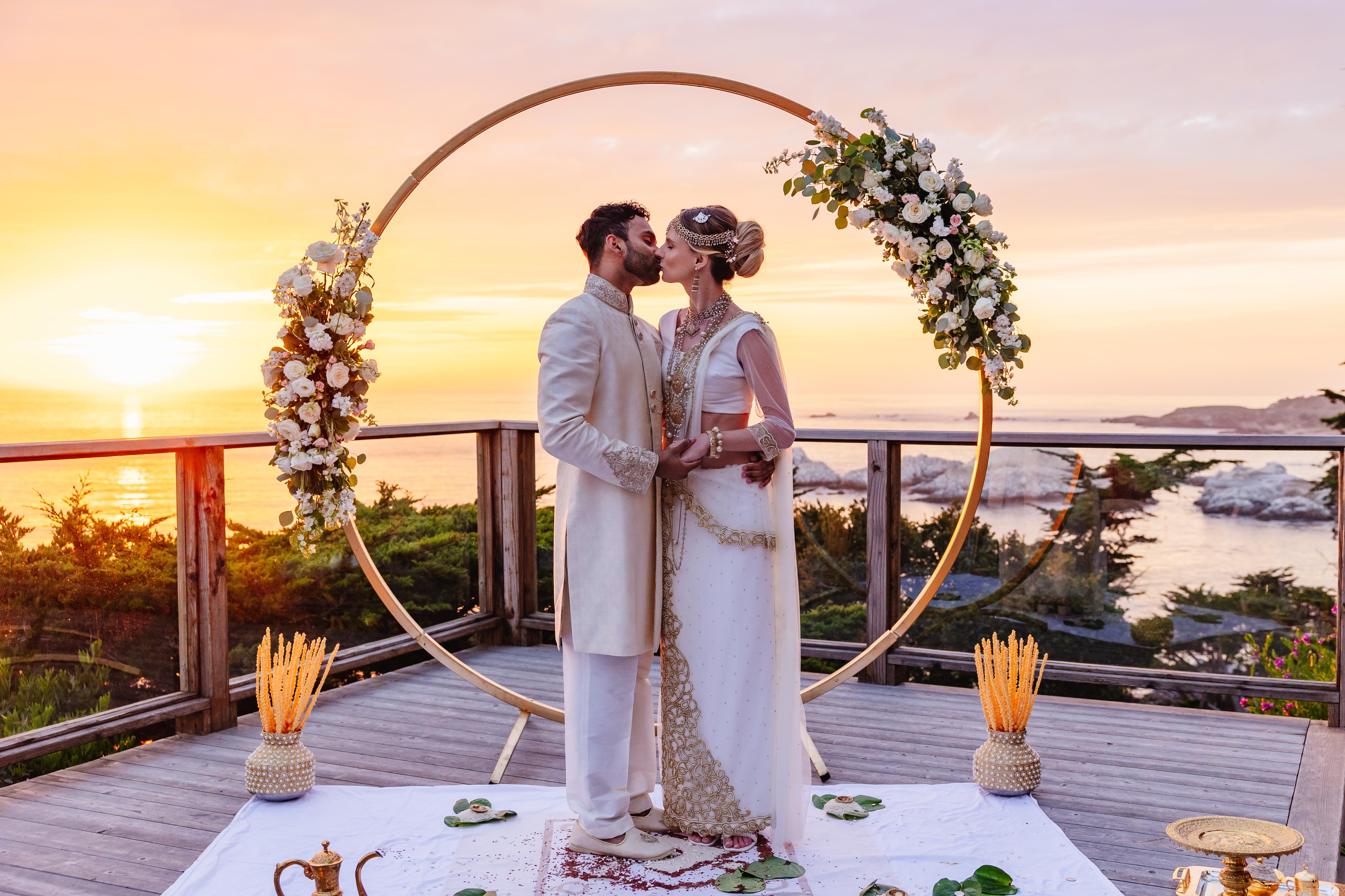 Couple in traditional attire kissing under a circular floral arch at sunset with an ocean view.