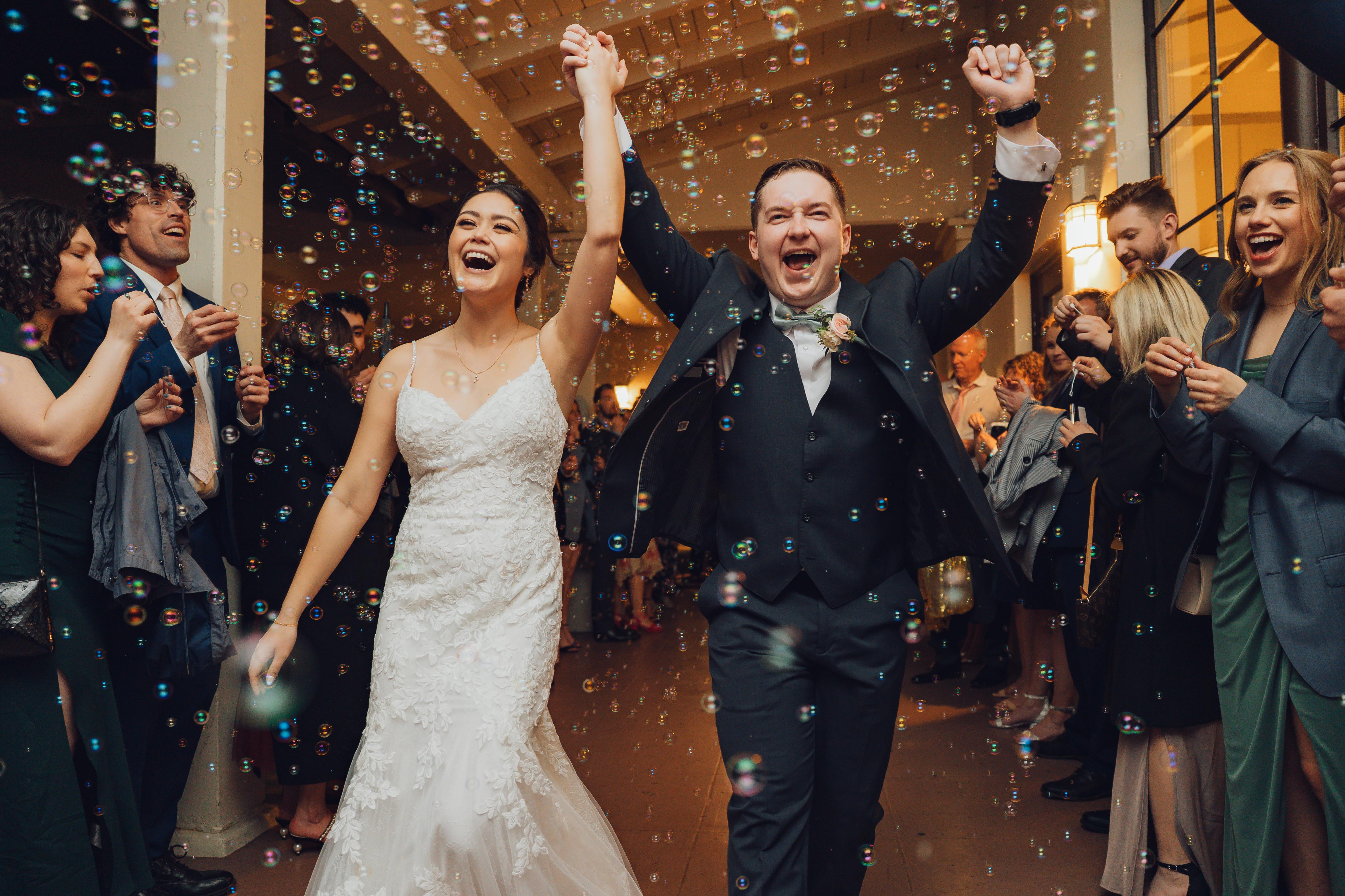 Bride and groom joyfully celebrating with raised hands surrounded by guests blowing bubbles.