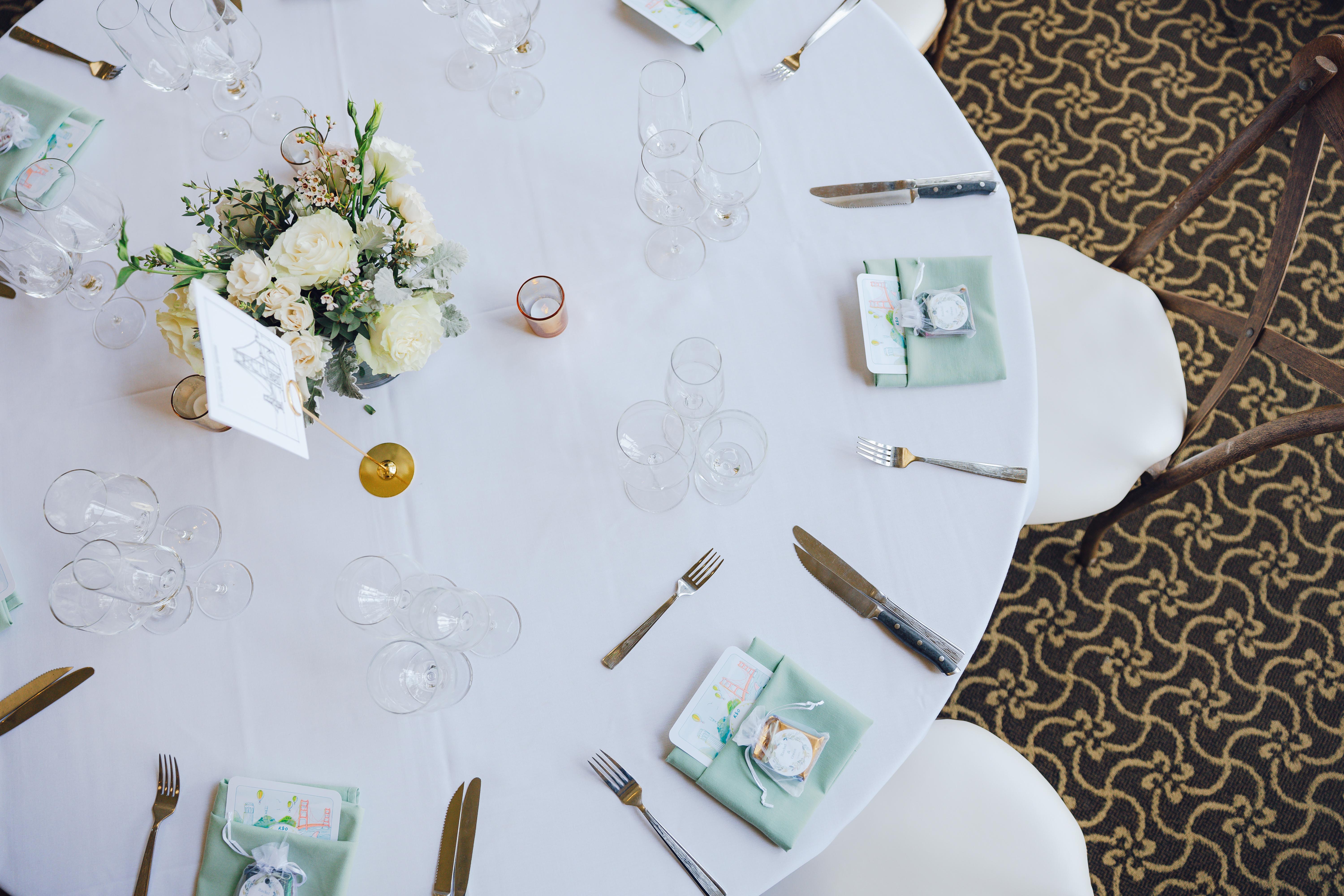 Round table with white tablecloth set for four, decorated with a floral centerpiece and place settings including green napkins, utensils, and glassware.