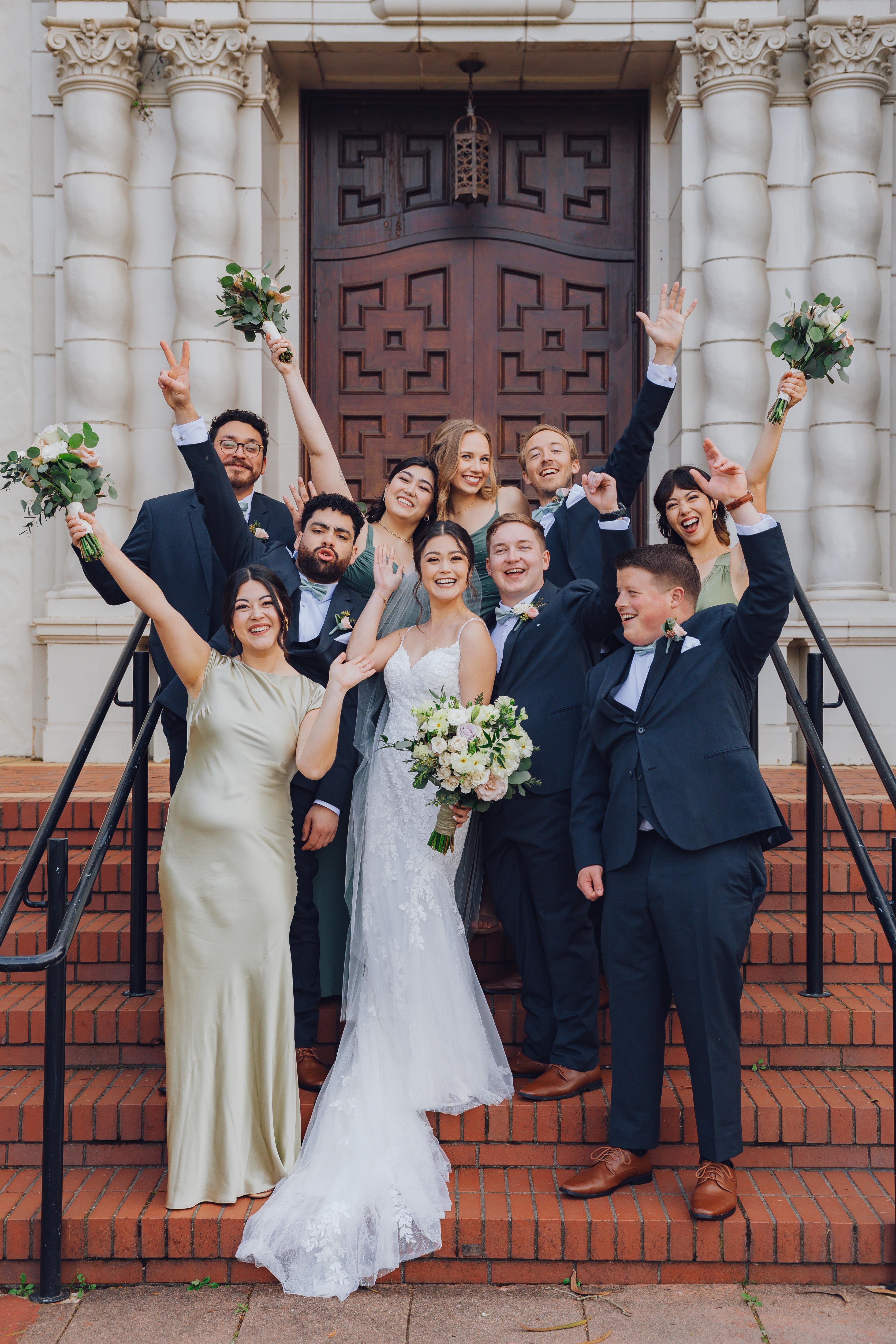 Bride in a white lace gown and groom in a dark suit surrounded by bridesmaids and groomsmen celebrating on outdoor steps.