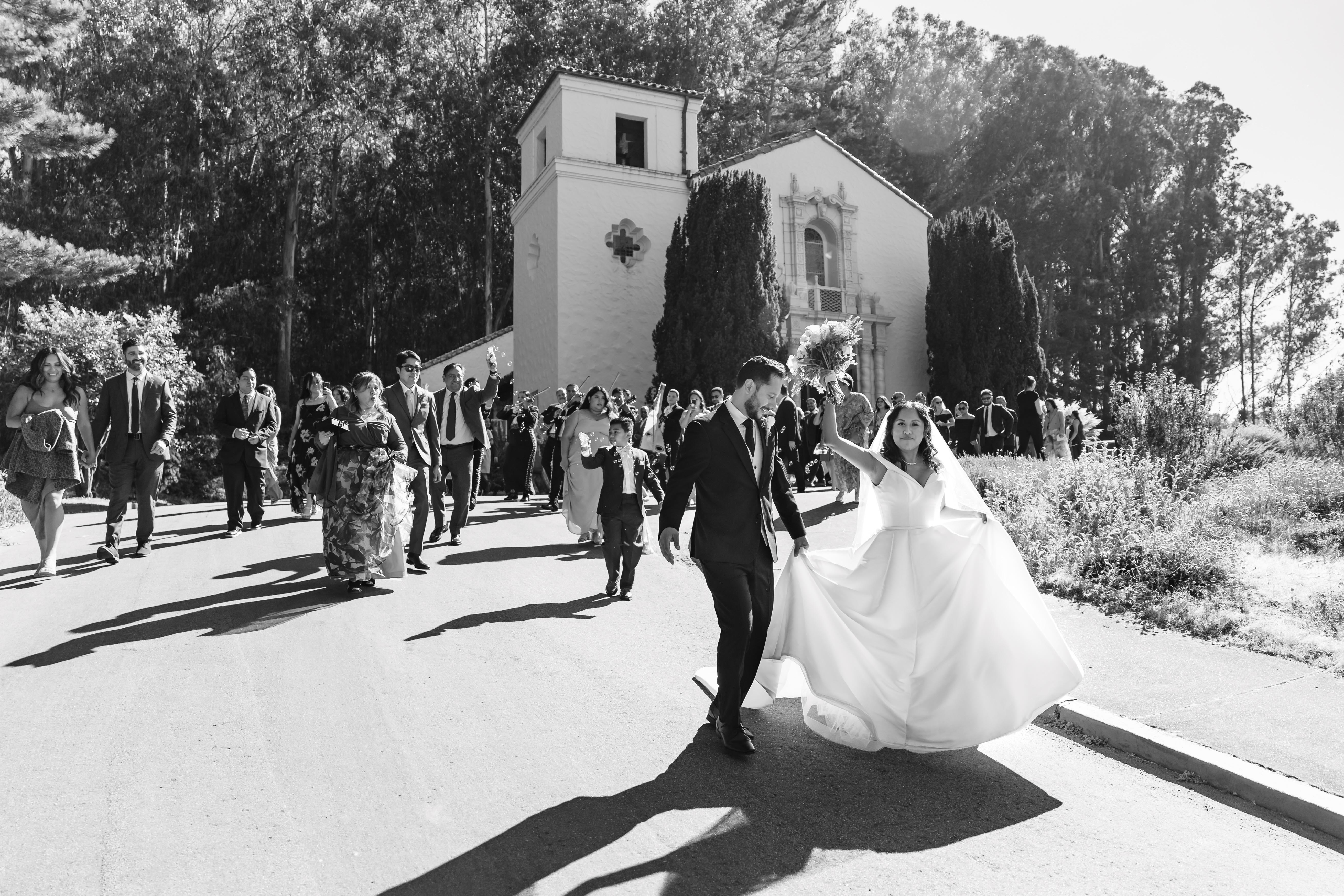 Bride and groom walking down a road with wedding guests behind them in front of a chapel surrounded by trees.