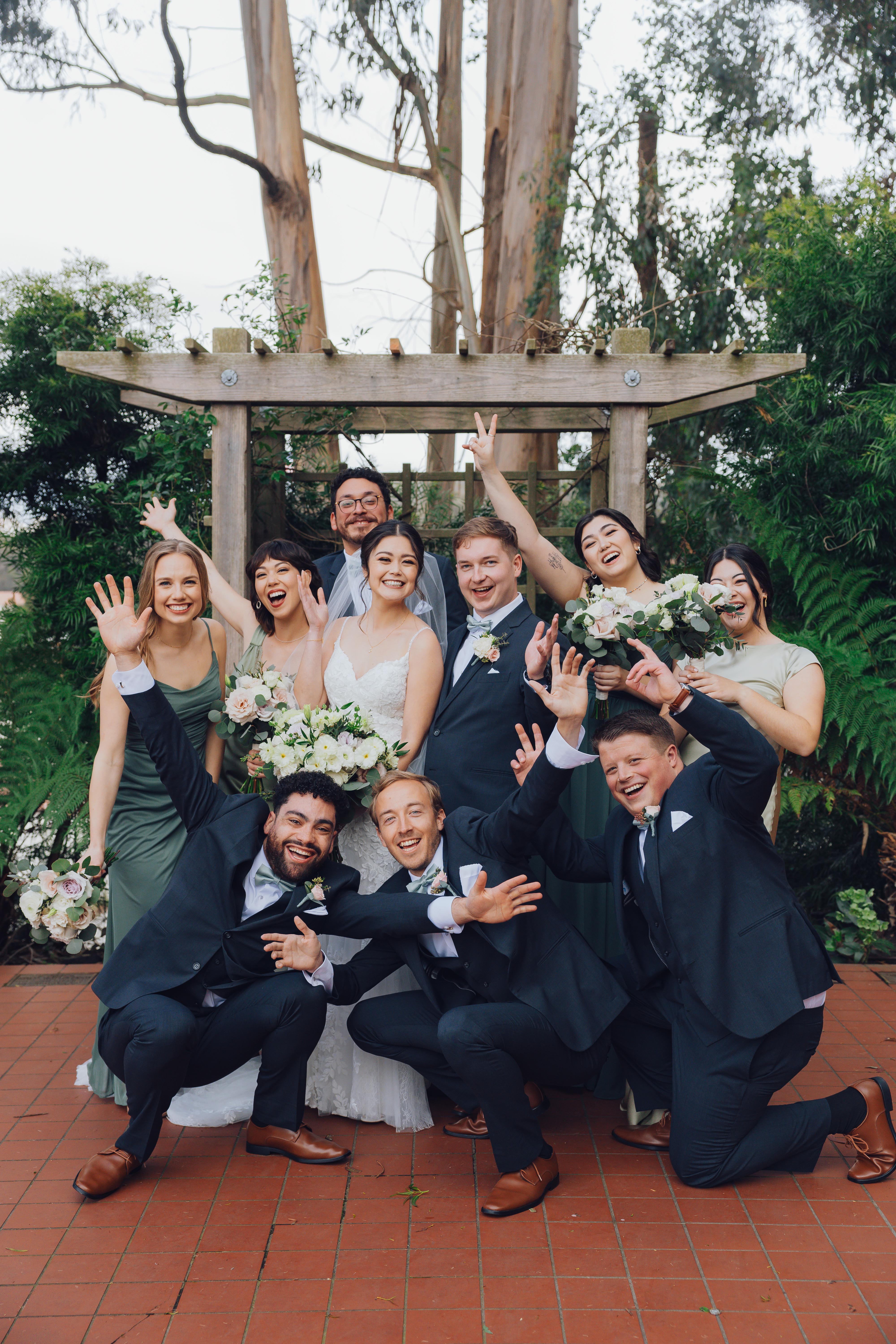 Bride and groom with bridesmaids and groomsmen posing happily under a wooden arbor outdoors.