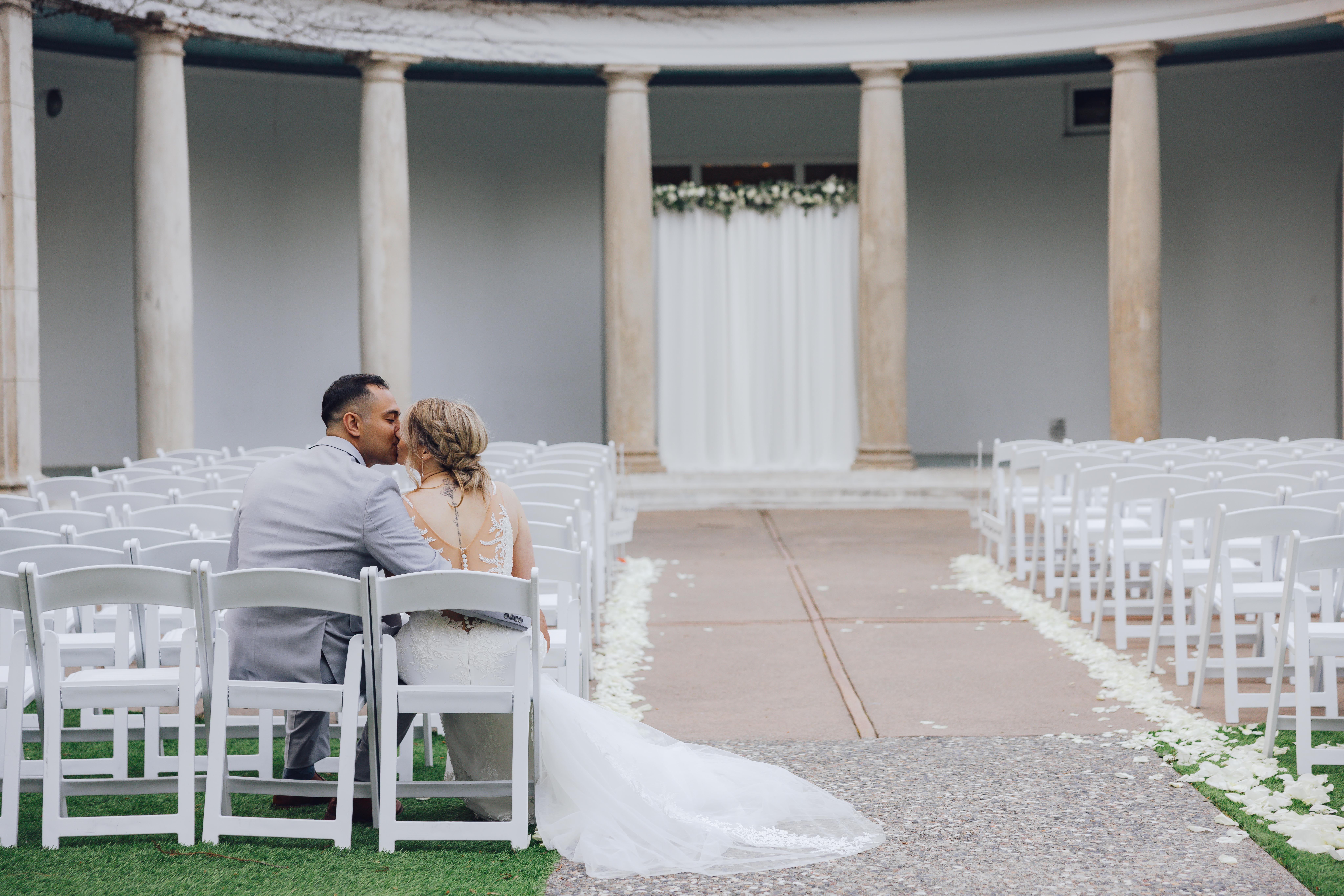 Bride and groom sitting and kissing on white chairs in an empty courtyard wedding venue with stone columns and floral decorations.