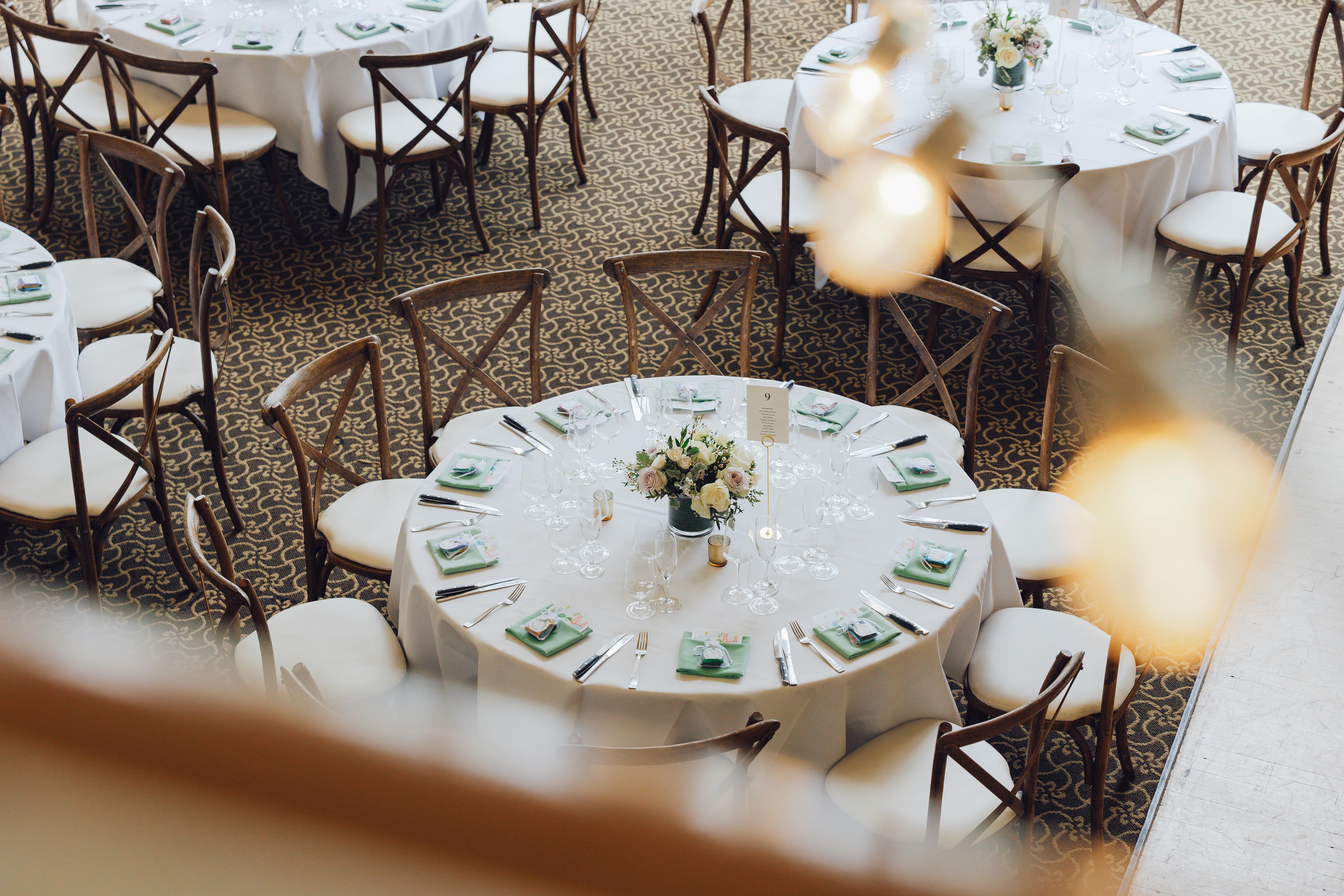 Round banquet tables with white tablecloths set with floral centerpieces, glassware, silverware, and mint green napkins, surrounded by wooden chairs on patterned carpet.