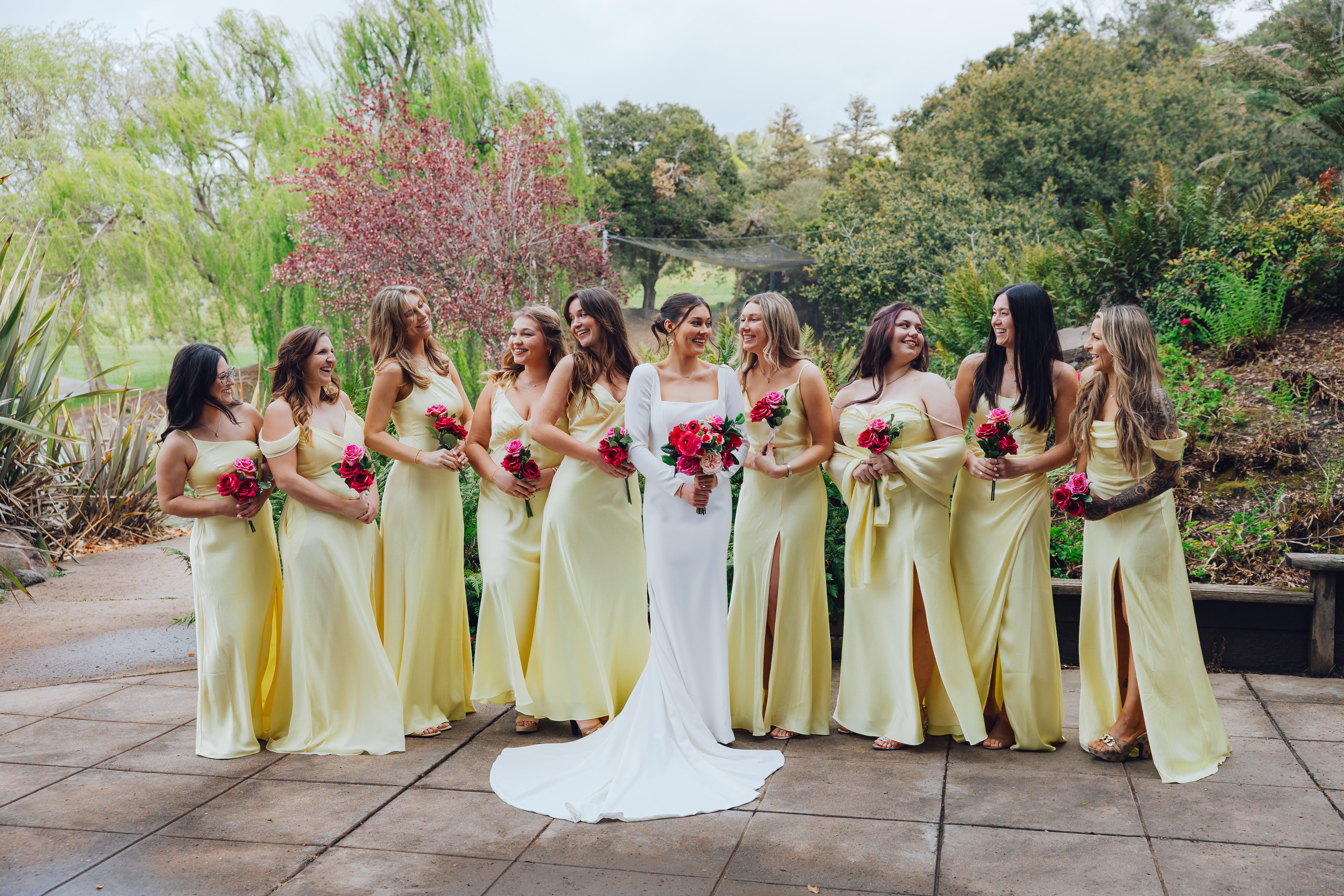 Bride in a white gown surrounded by bridesmaids in yellow dresses holding pink roses, standing outdoors with greenery.