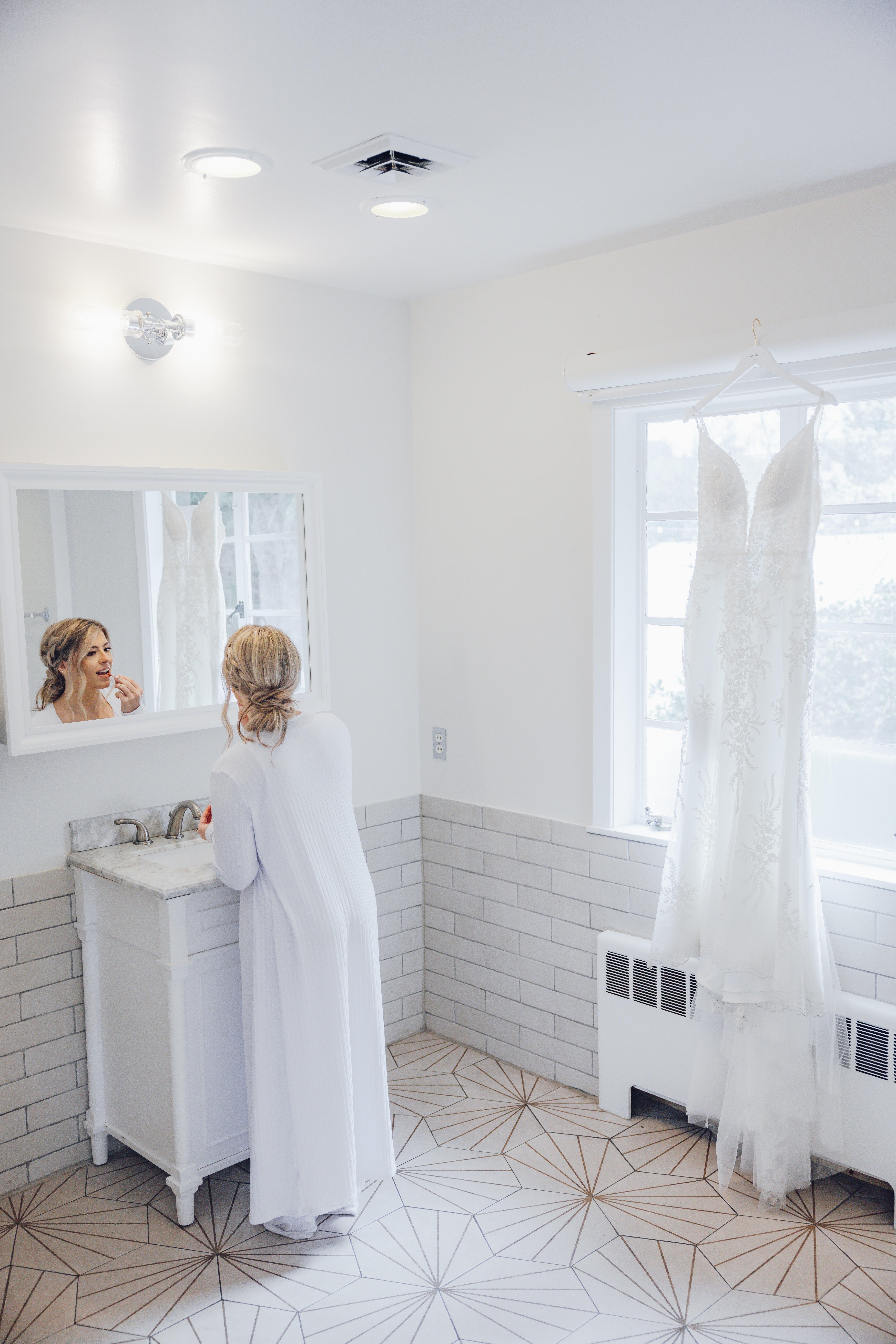 Woman in white robe applying lipstick while looking in bathroom mirror with a white wedding dress hanging by the window.