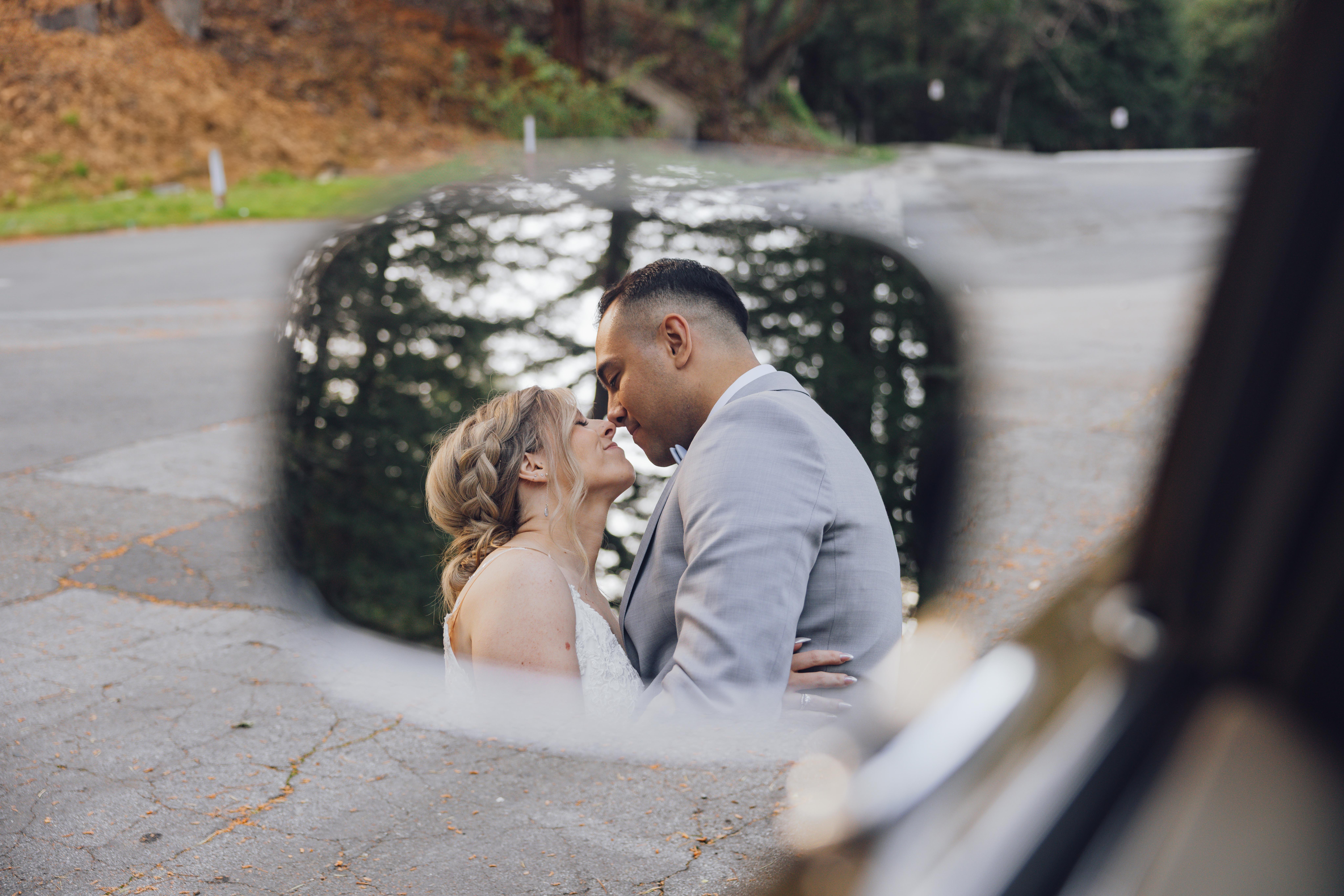 Bride and groom embracing and about to kiss, reflected in a car's side mirror.
