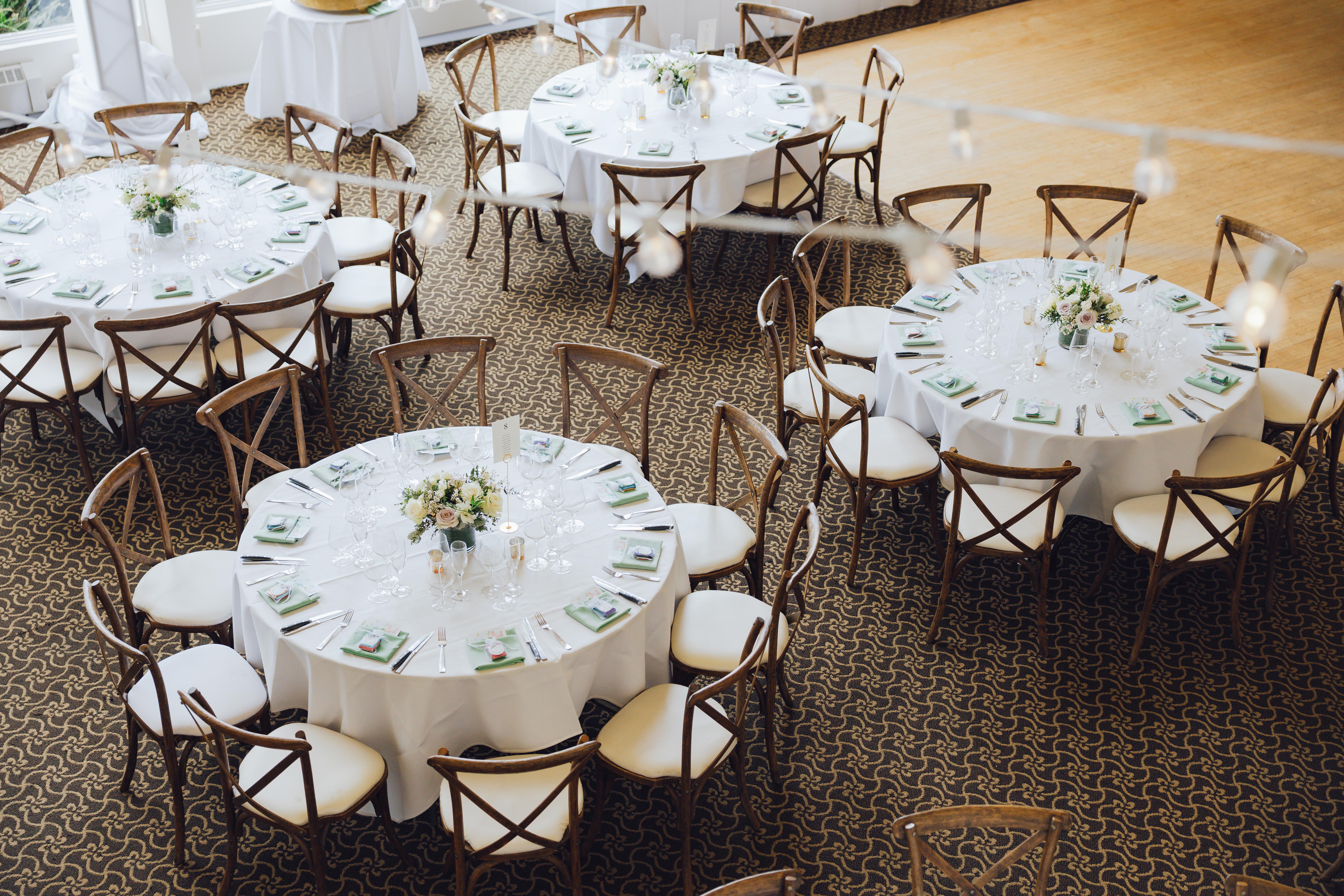 Round banquet tables set with white tablecloths, floral centerpieces, glassware, cutlery, and napkins, surrounded by wooden chairs with white cushions.