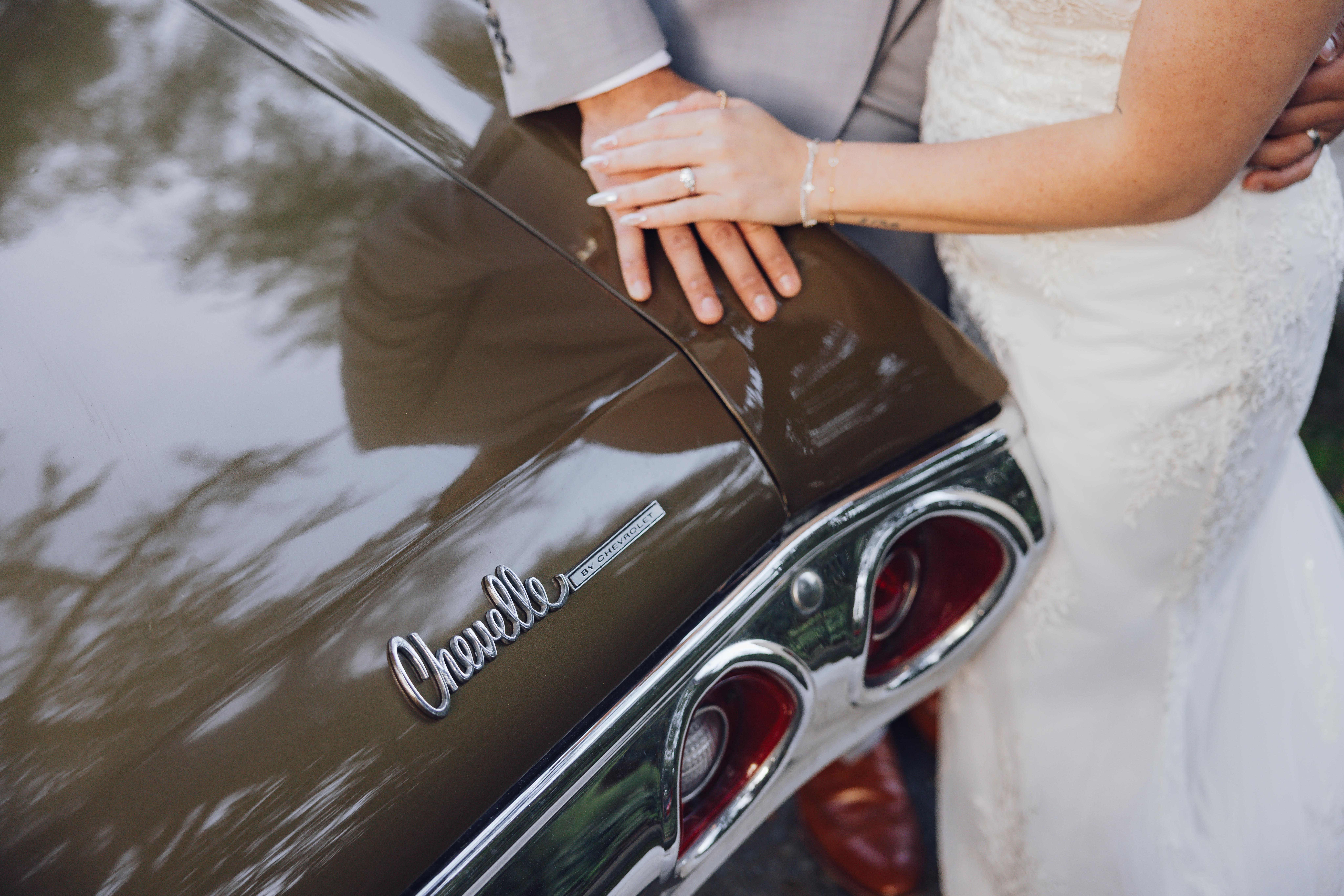 Close-up of a bride and groom's hands resting on the trunk of a brown Chevrolet Chevelle car, showing the car's badge and rear lights.