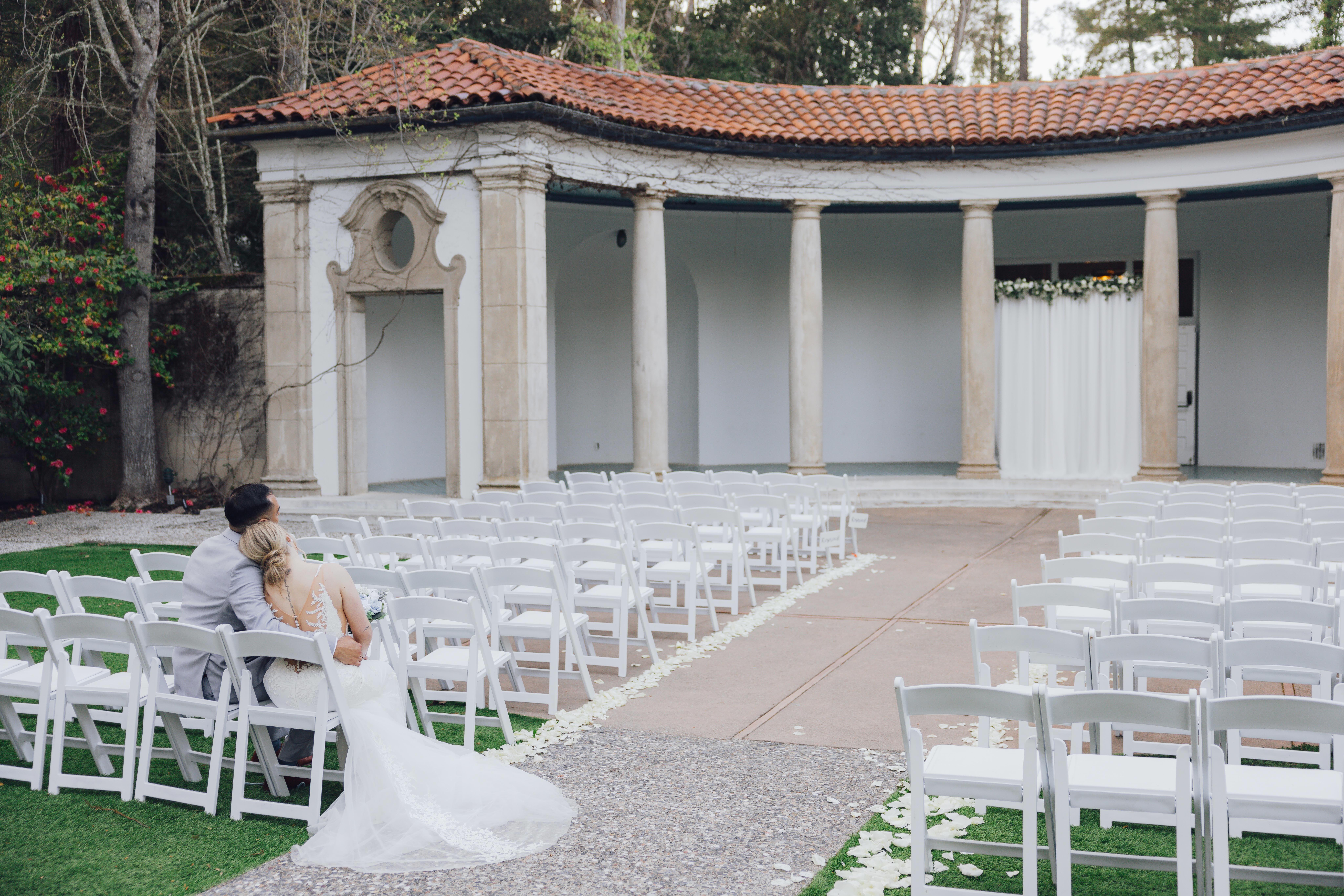 Bride and groom sitting together on white chairs in an outdoor wedding venue with stone columns and flower petals along the aisle.