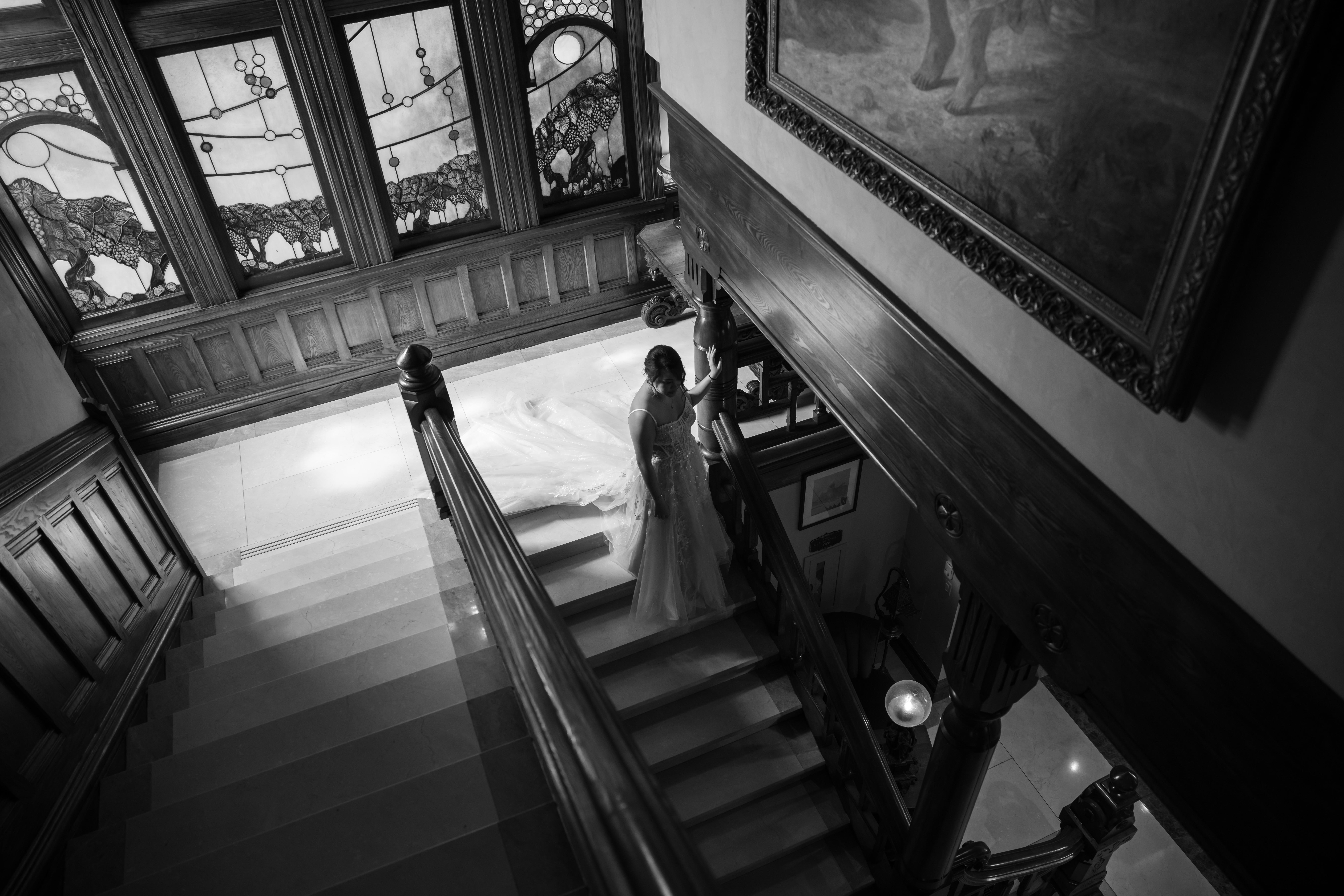 Bride in a wedding gown descending an ornate wooden staircase with stained glass windows and a large framed painting nearby.