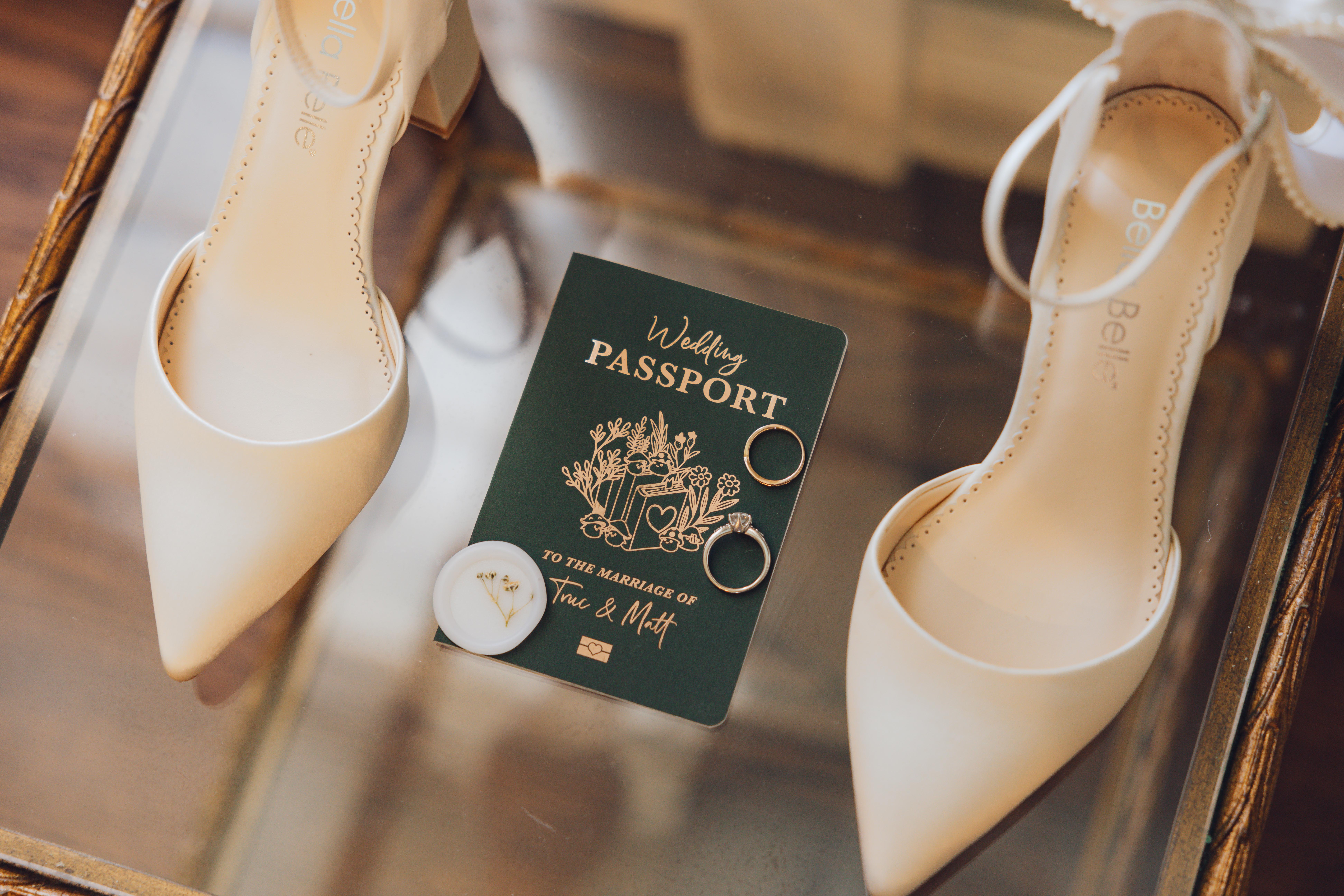 Pair of white bridal shoes beside a green wedding passport booklet with two rings and a small white wax seal on a glass surface.