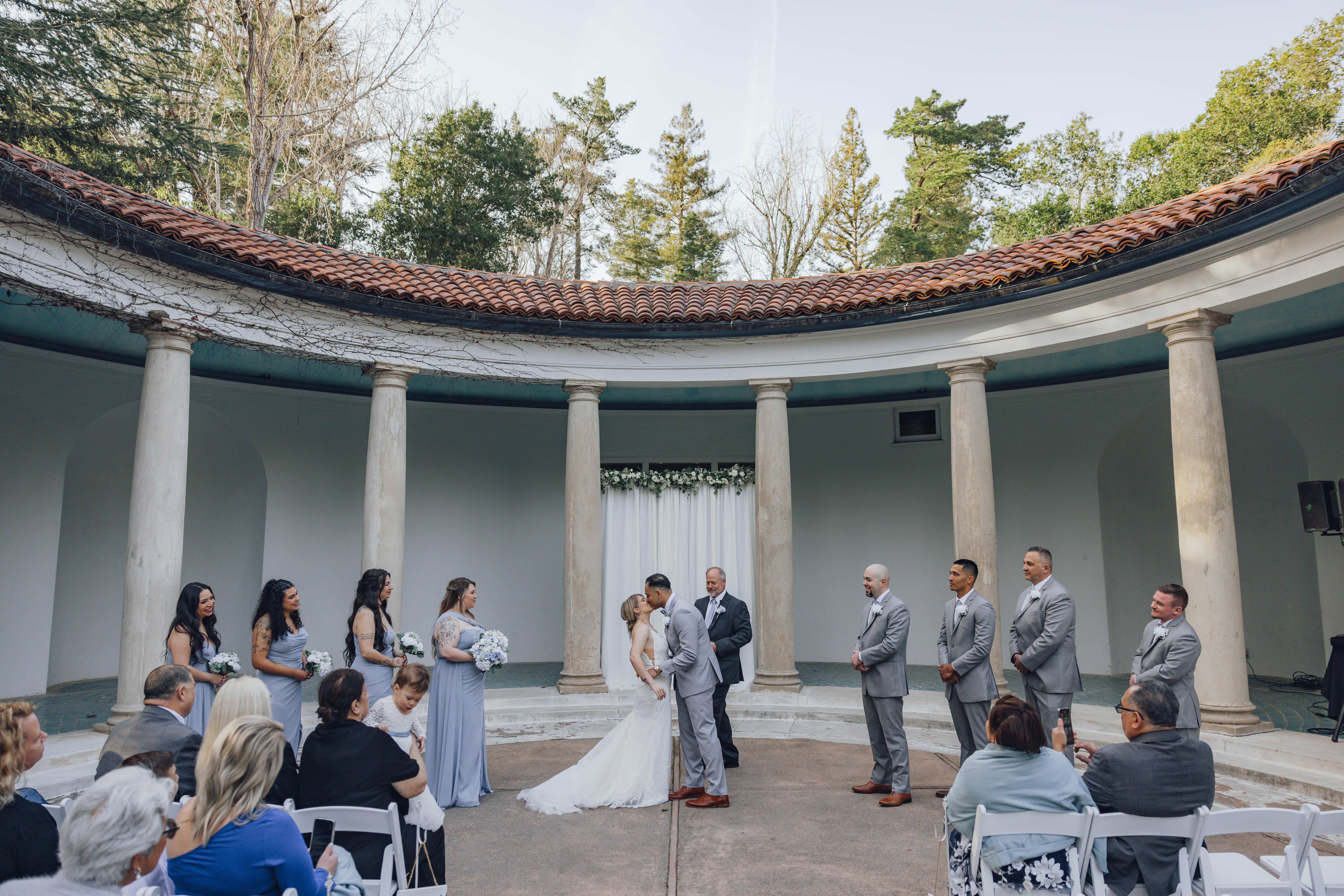 Bride and groom kissing under a columned circular structure during an outdoor wedding ceremony with bridesmaids and groomsmen standing on either side.