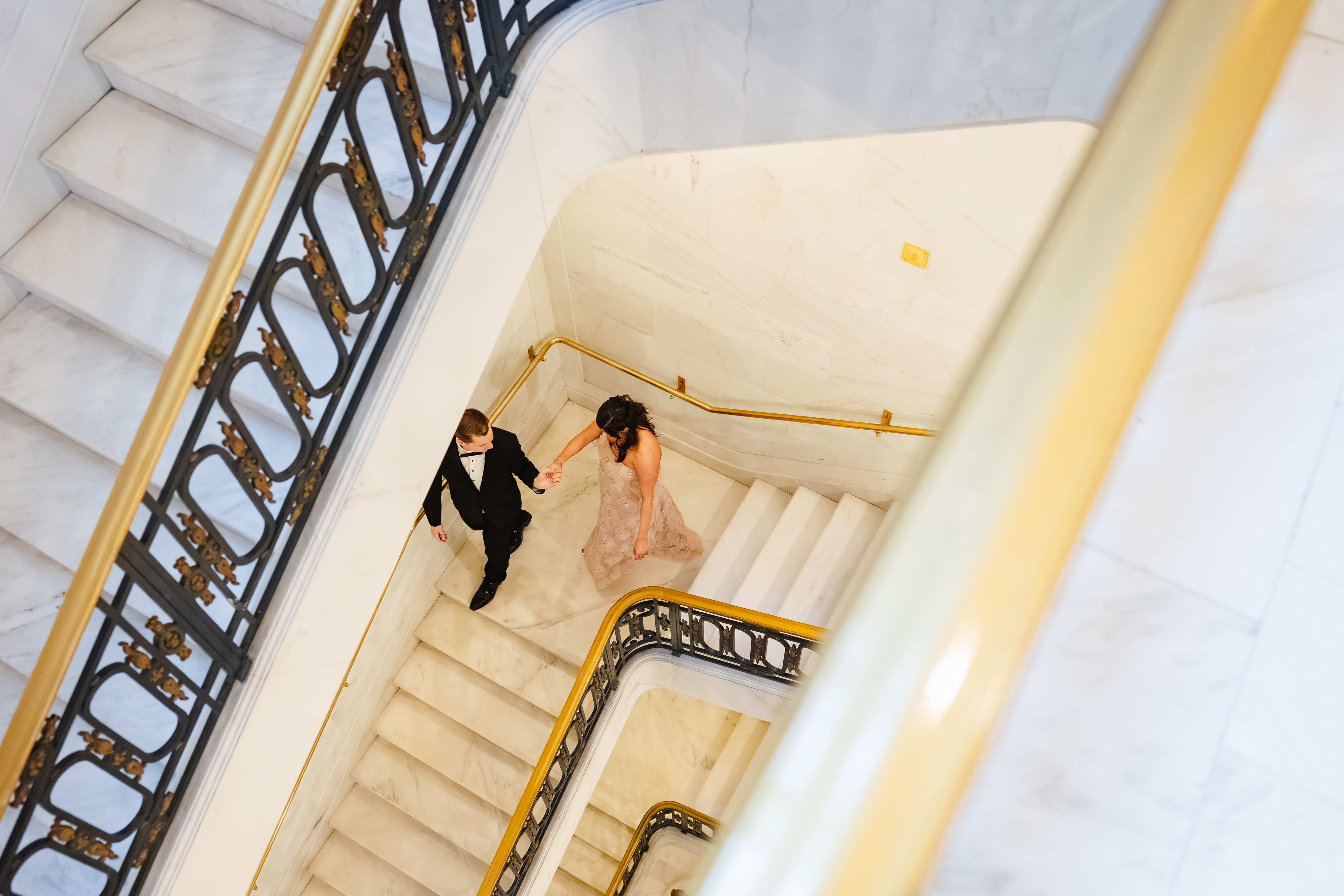 Overhead view of a couple holding hands as they walk up a white marble staircase with gold and black railings.
