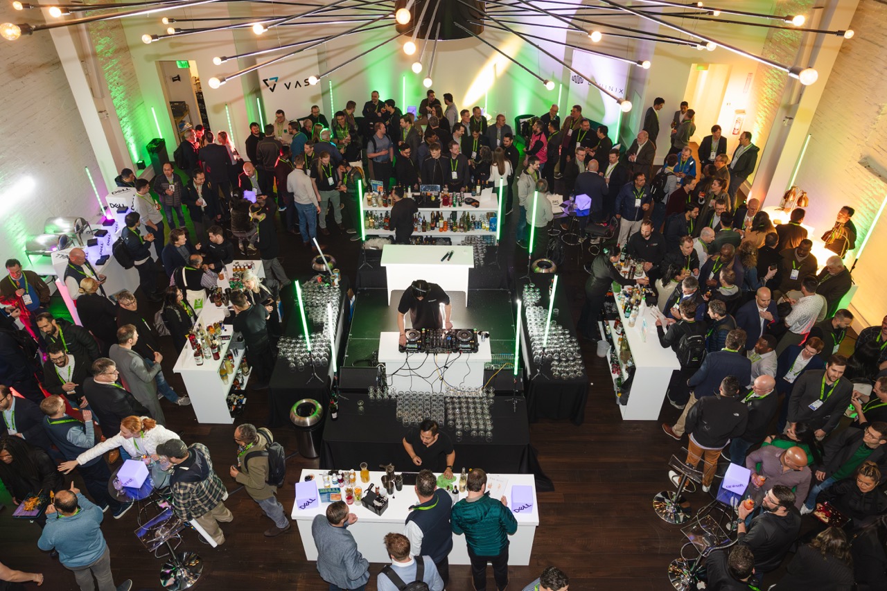 Overhead view of a crowded indoor event with people mingling around white bars and a DJ playing music at the center.