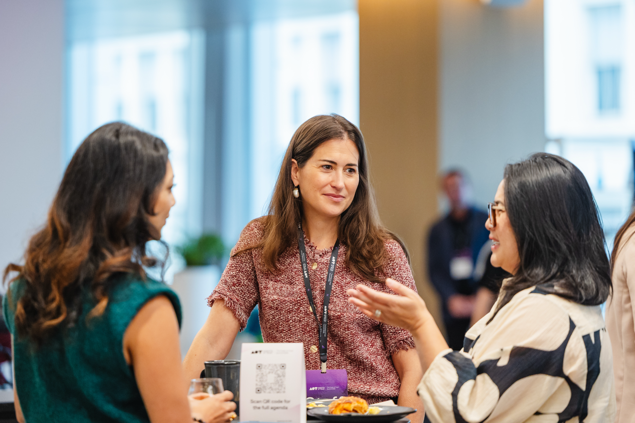 Three women engaged in conversation at a conference with food and drink on the table.