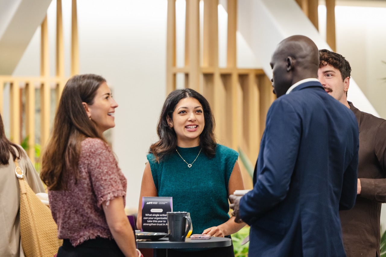 Four colleagues having a conversation around a small round table in a modern office setting.