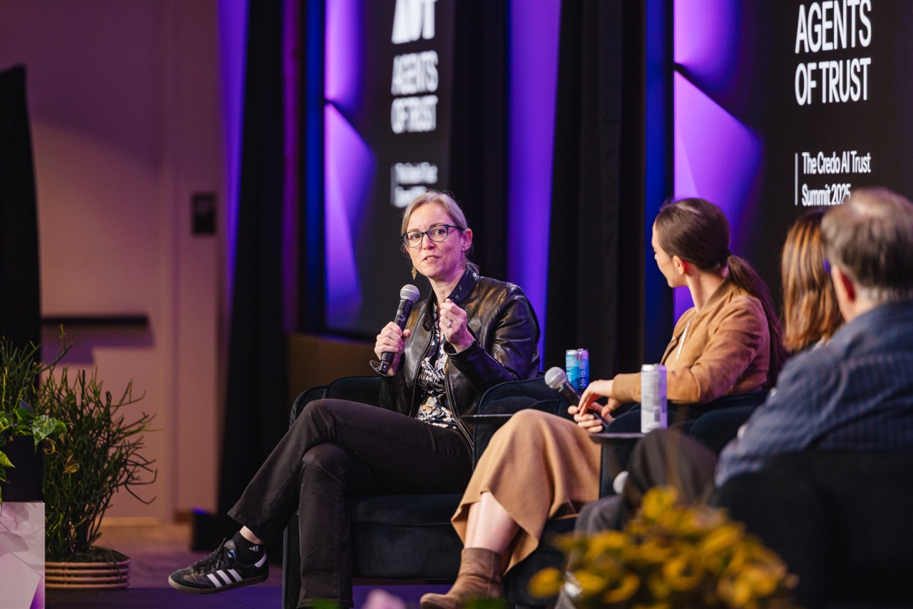 Panel discussion at Agents of Trust event with a woman speaking into a microphone and others seated, with purple-lit background.