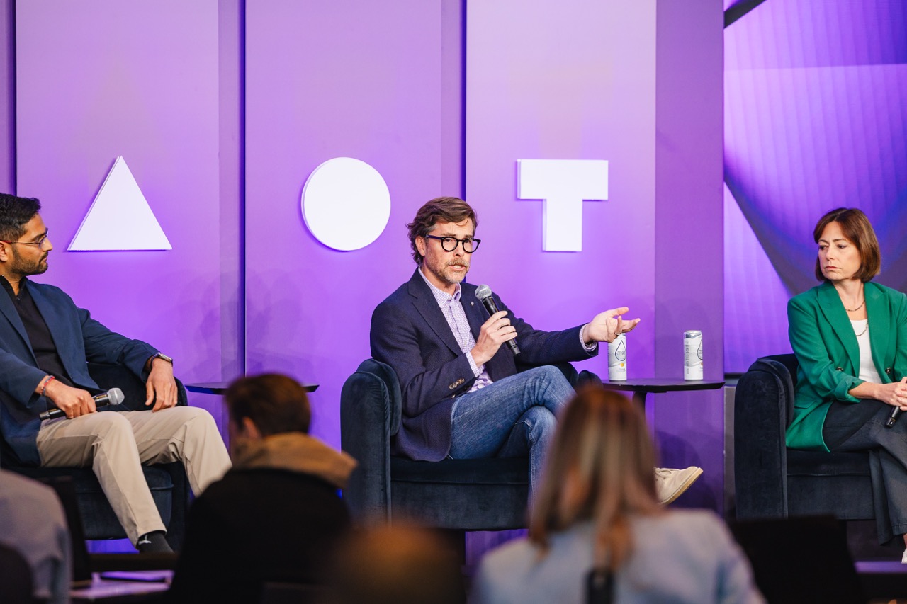Three panelists seated and speaking on stage with purple background featuring shapes triangle, circle, and T.