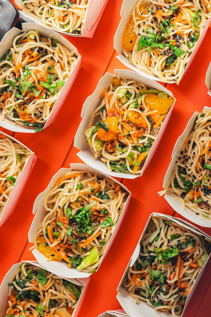 Multiple takeout boxes filled with stir-fried noodles mixed with vegetables, garnished with chopped herbs and black sesame seeds on an orange surface.