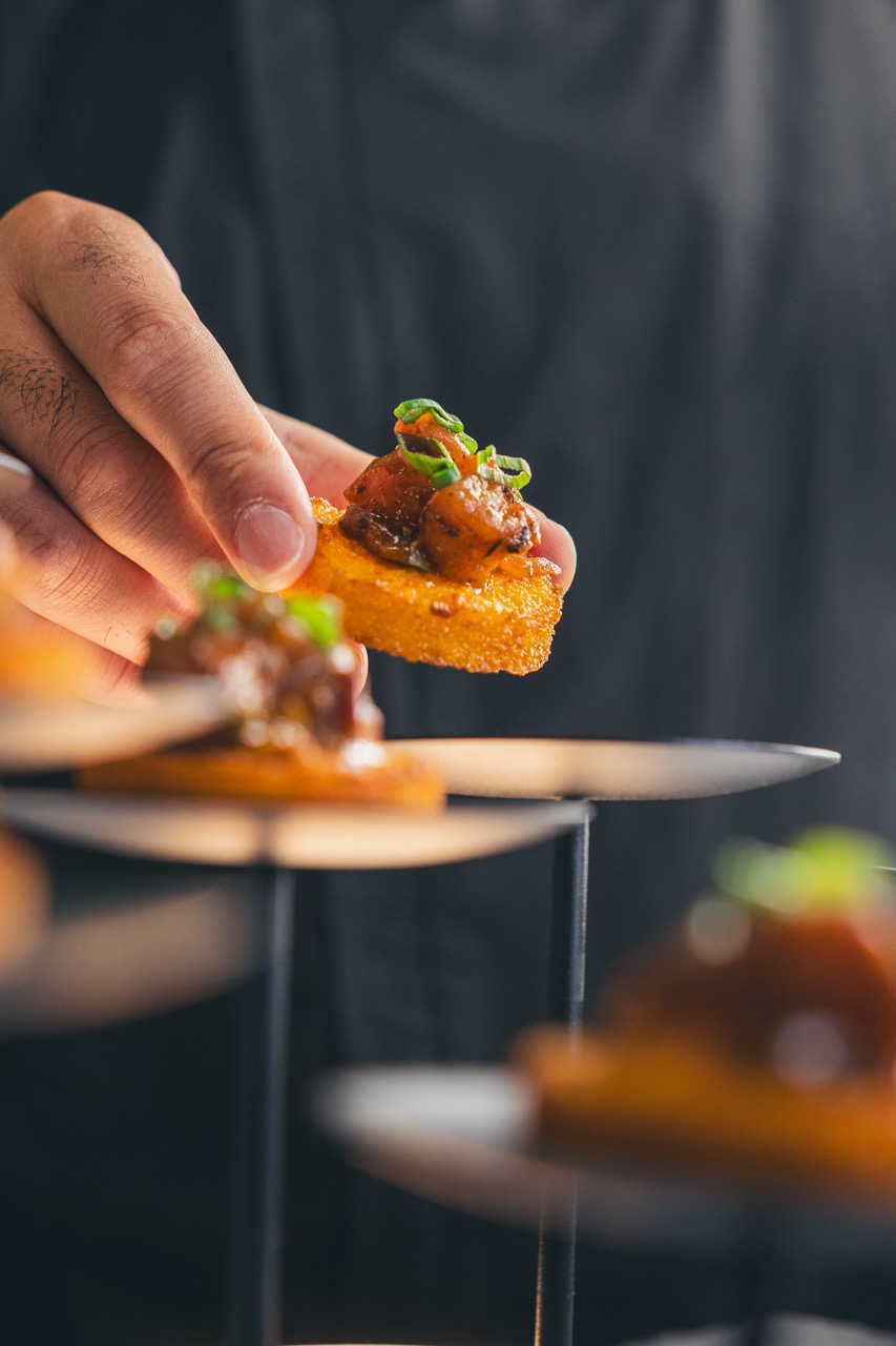 Hand picking up a bite-sized crispy appetizer topped with seasoned meat and garnished with chopped green herbs.