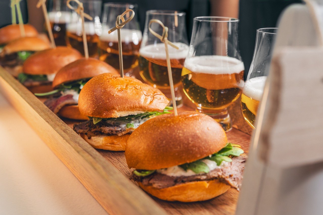 A wooden tray with four small sandwiches held by decorative toothpicks, accompanied by four glasses of beer.