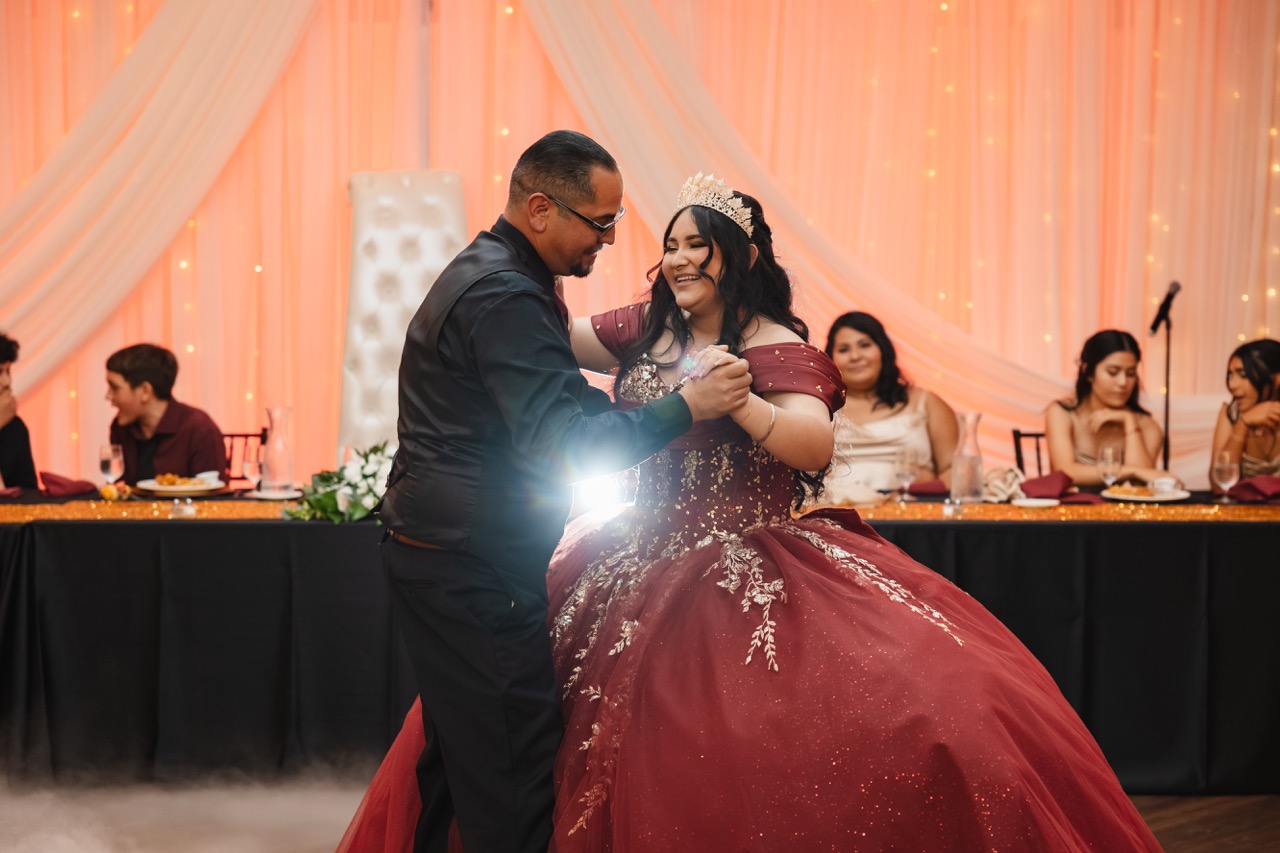 Young woman in a burgundy ball gown and tiara dancing closely with a man in black attire at a decorated event with seated guests in the background.