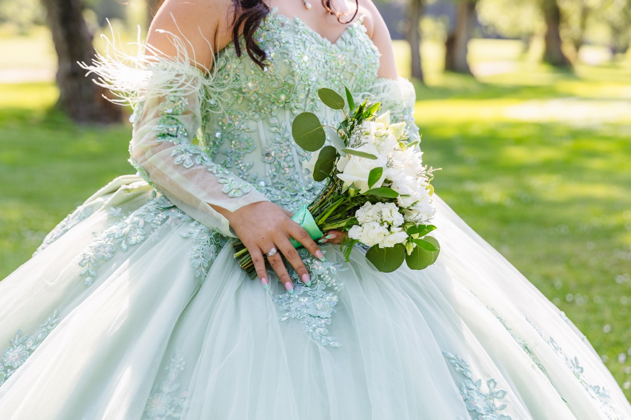 Close-up of a person in an ornate light mint green quinceañera dress holding a bouquet of white flowers and greenery outdoors.