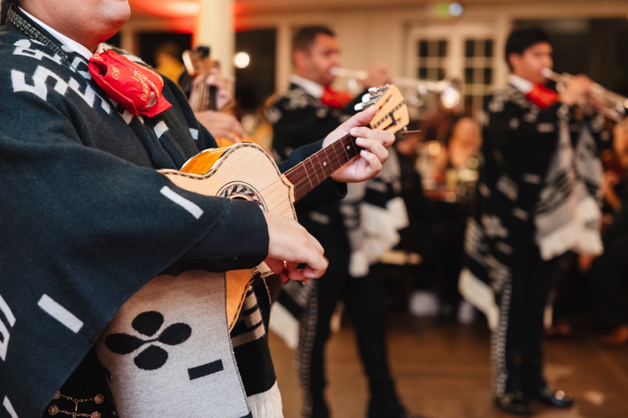 Mariachi musicians wearing traditional outfits and red bow ties playing string and brass instruments indoors.