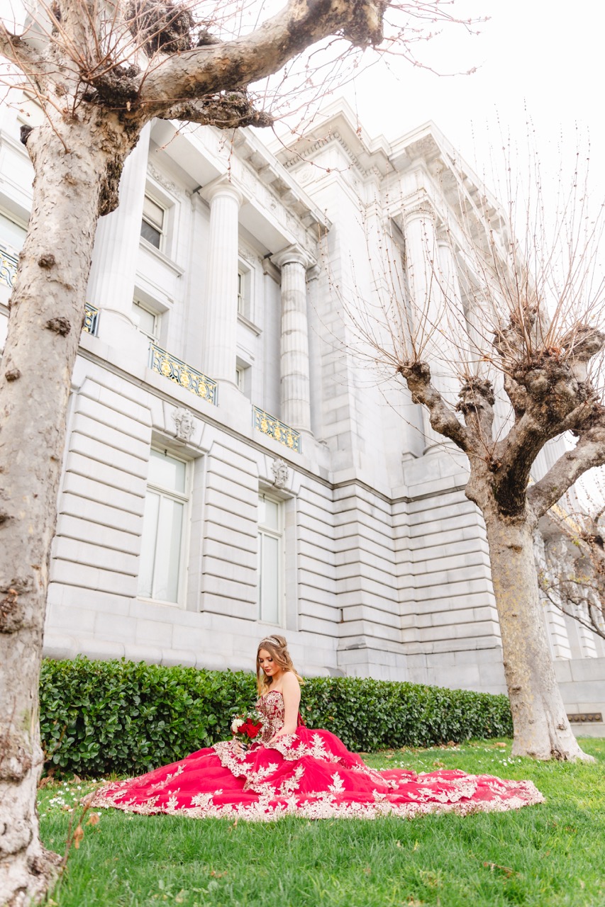 Woman in a red strapless gown with gold embroidery sitting on grass in front of a neoclassical stone building with columns and leafless trees.