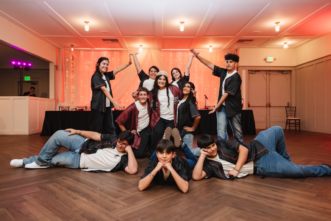 Group of nine smiling teenagers posing indoors with one girl wearing a tiara and maroon shirt in the center.