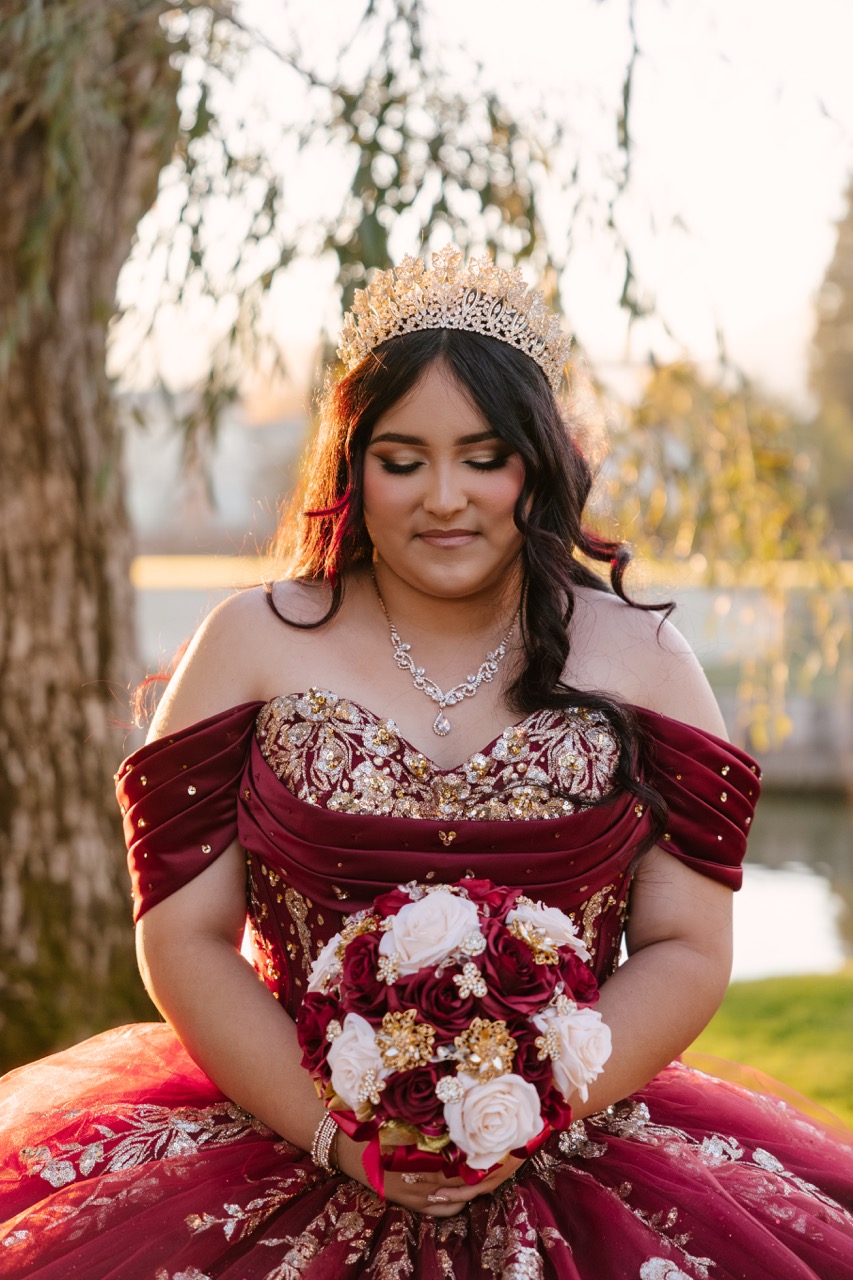 Young woman in a burgundy and gold off-shoulder gown with a jeweled tiara holding a bouquet of red and white roses outdoors.