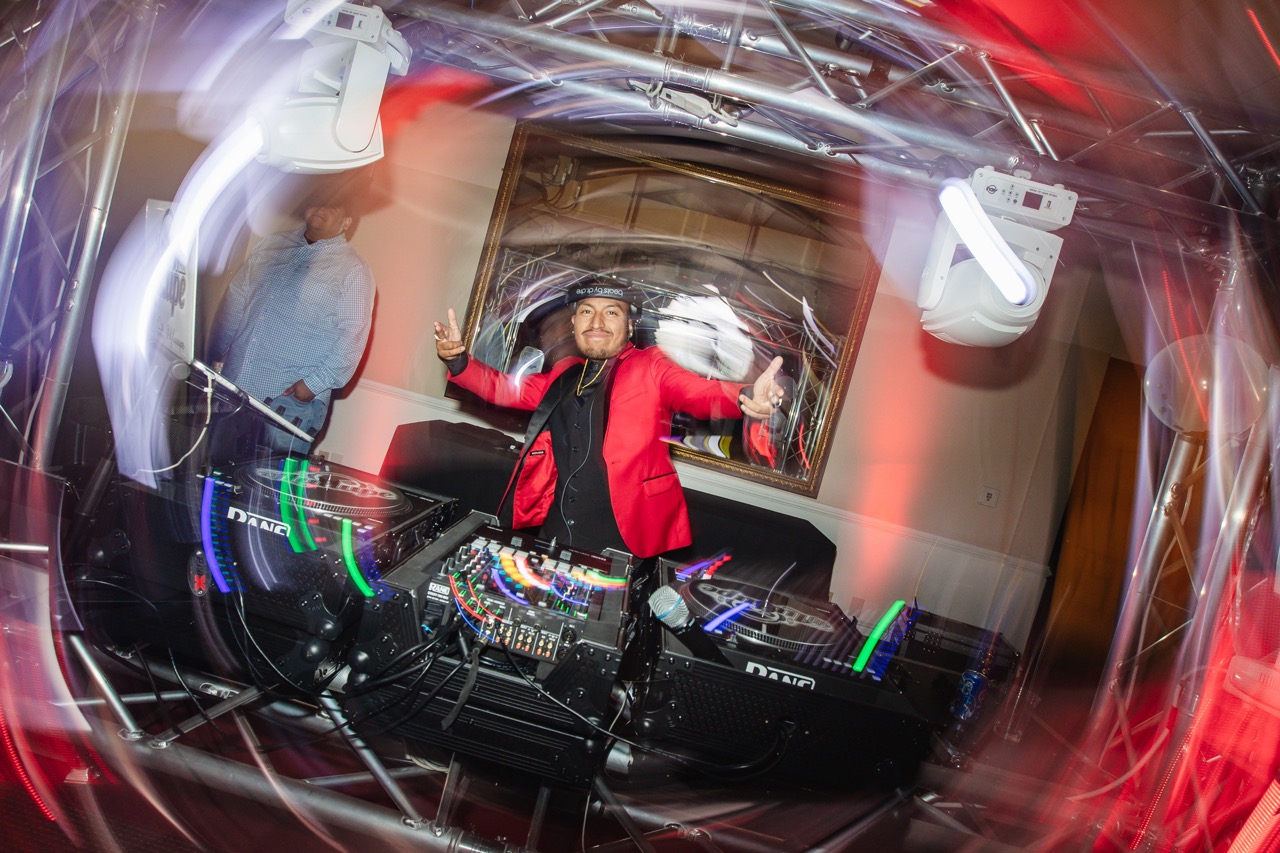 DJ wearing a red jacket and black cap stands behind a mixing console with colorful light trails around him.