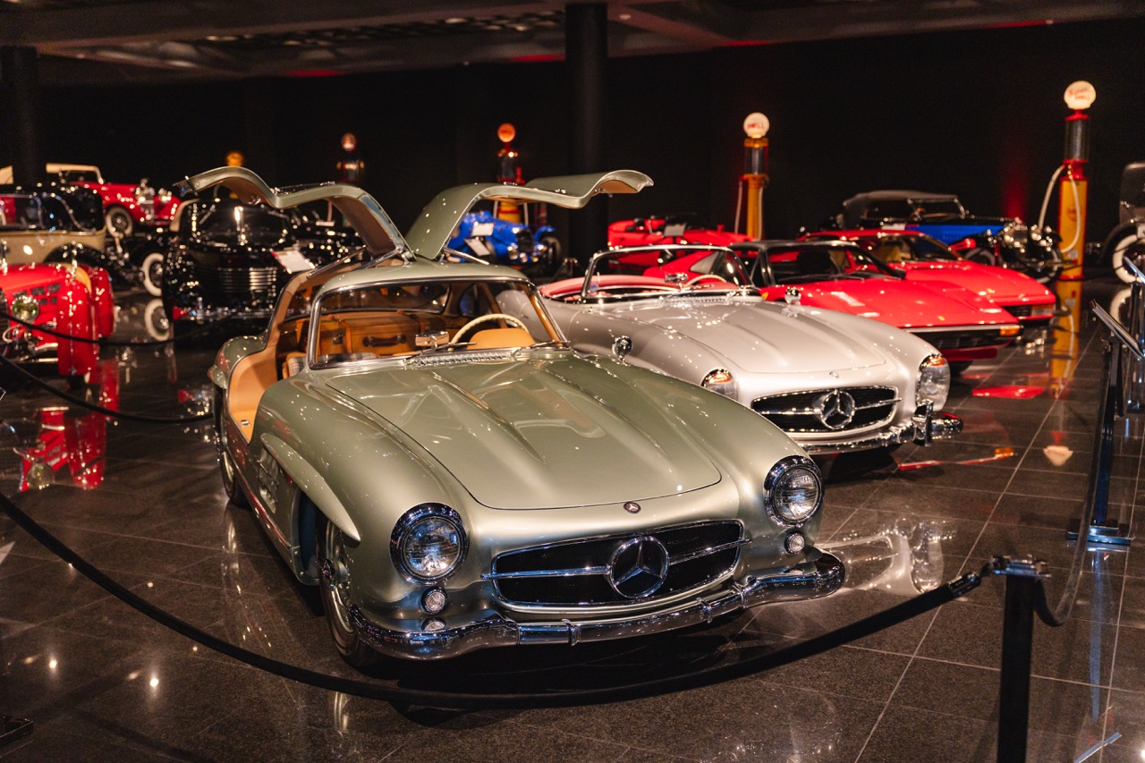 Classic Mercedes-Benz cars in a museum, featuring a silver model with gull-wing doors open in the foreground.