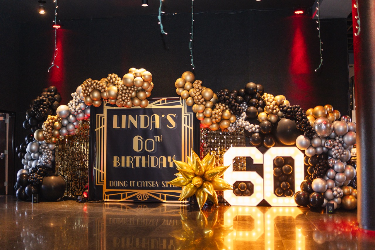 Elegant Gatsby-themed birthday decoration with gold, black, and silver balloons, a large illuminated '60', and a sign that reads 'Linda's 60th Birthday'.