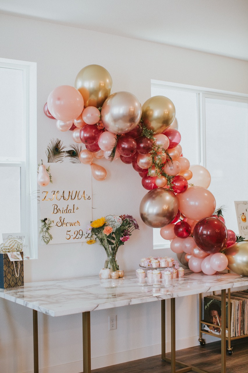 Decorated bridal shower table with pink, gold, and red balloons, a vase with flowers, cupcakes, and a sign reading 'Zhanna's Bridal Shower 5-29'.