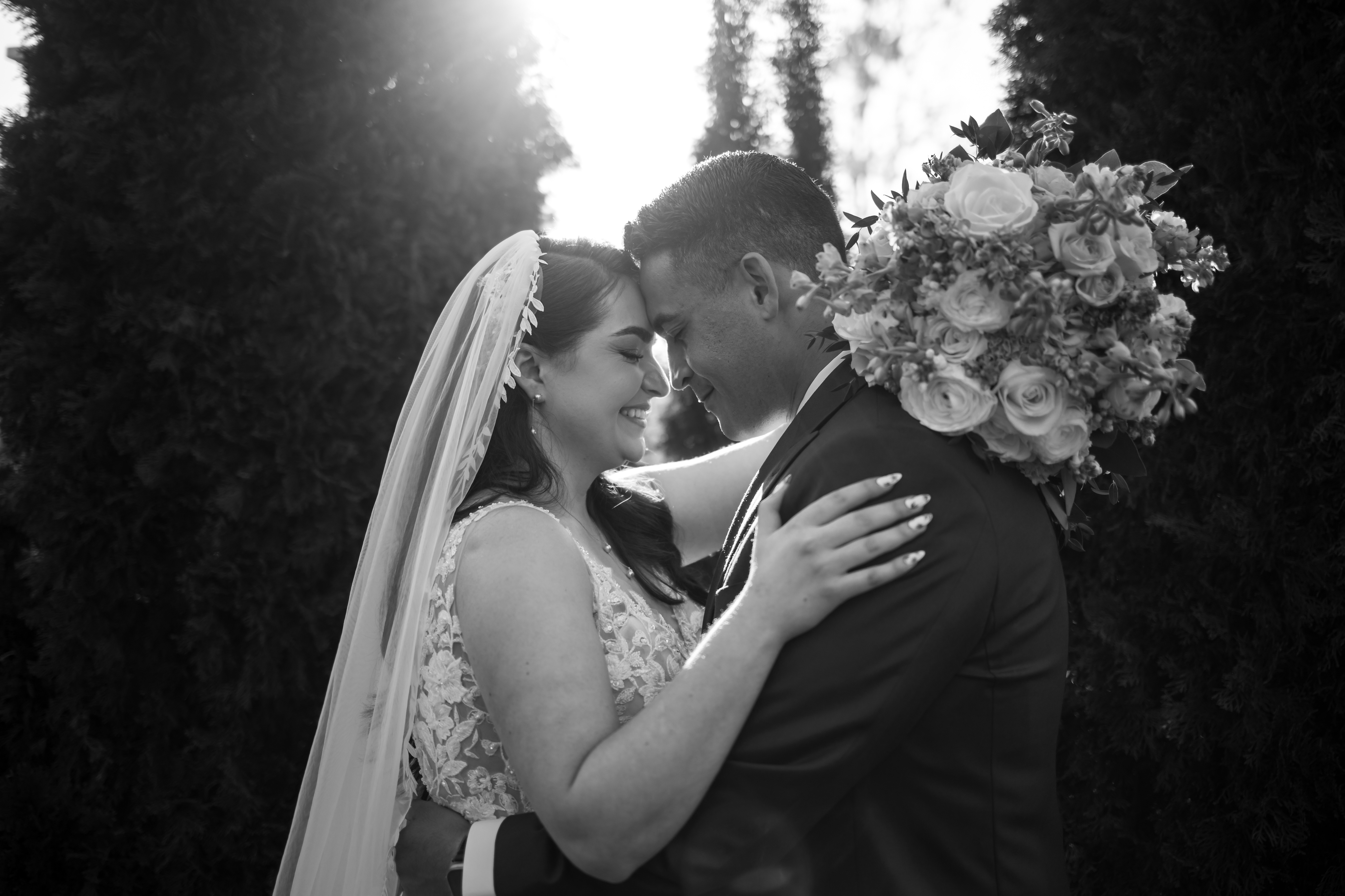 Black and white photo of a bride and groom embracing with foreheads touching, the bride wearing a lace dress and veil, and the groom holding a bouquet of roses.
