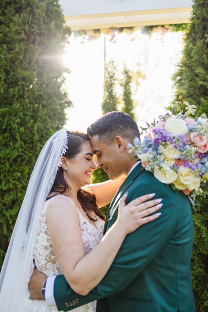 Bride and groom in a close embrace outdoors, the bride holding a colorful bouquet and both smiling with foreheads touching.