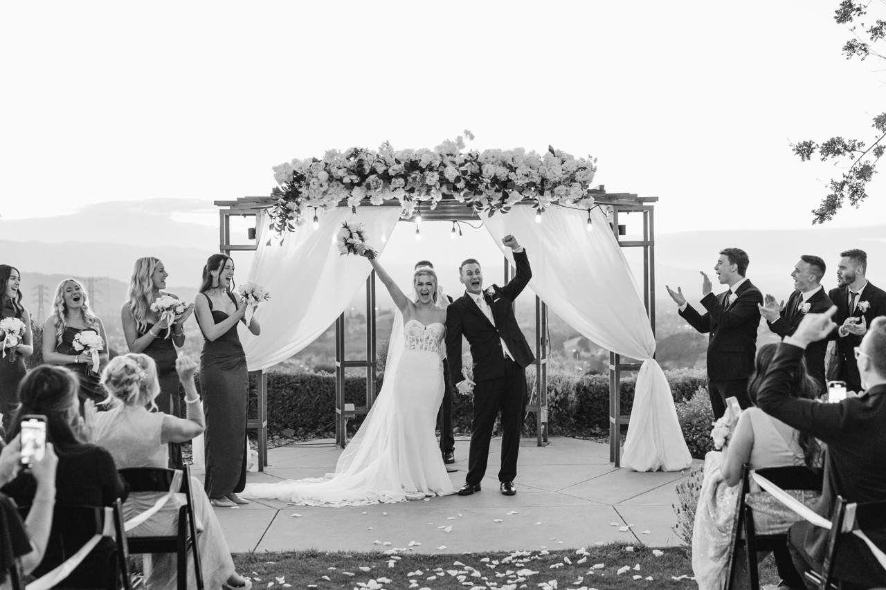 Bride and groom celebrating under a floral wedding arch with guests and bridal party clapping on either side.