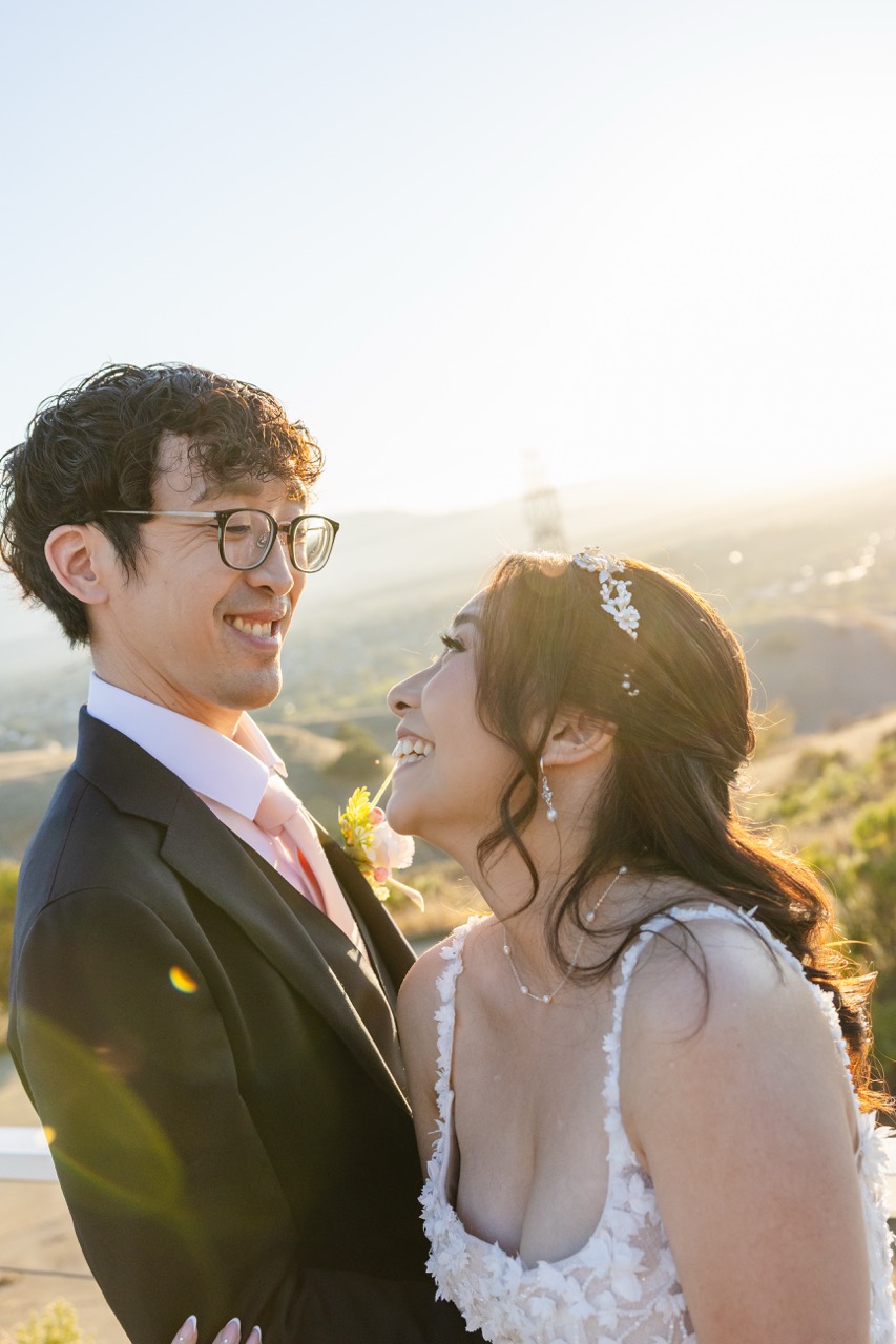 Bride and groom smiling and looking at each other outdoors at sunset, with the bride in a white floral dress and the groom in a black suit with glasses.