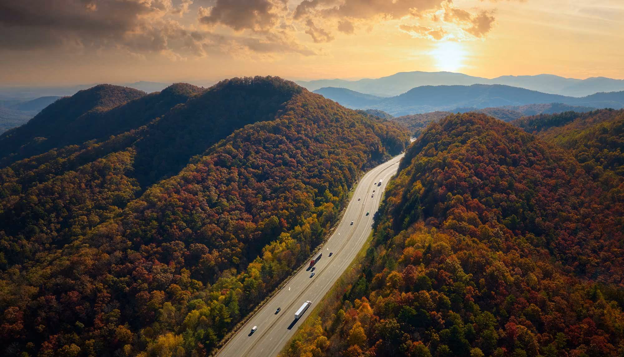 Aerial view of an interstate winding through forested mountains at sunset with trucks